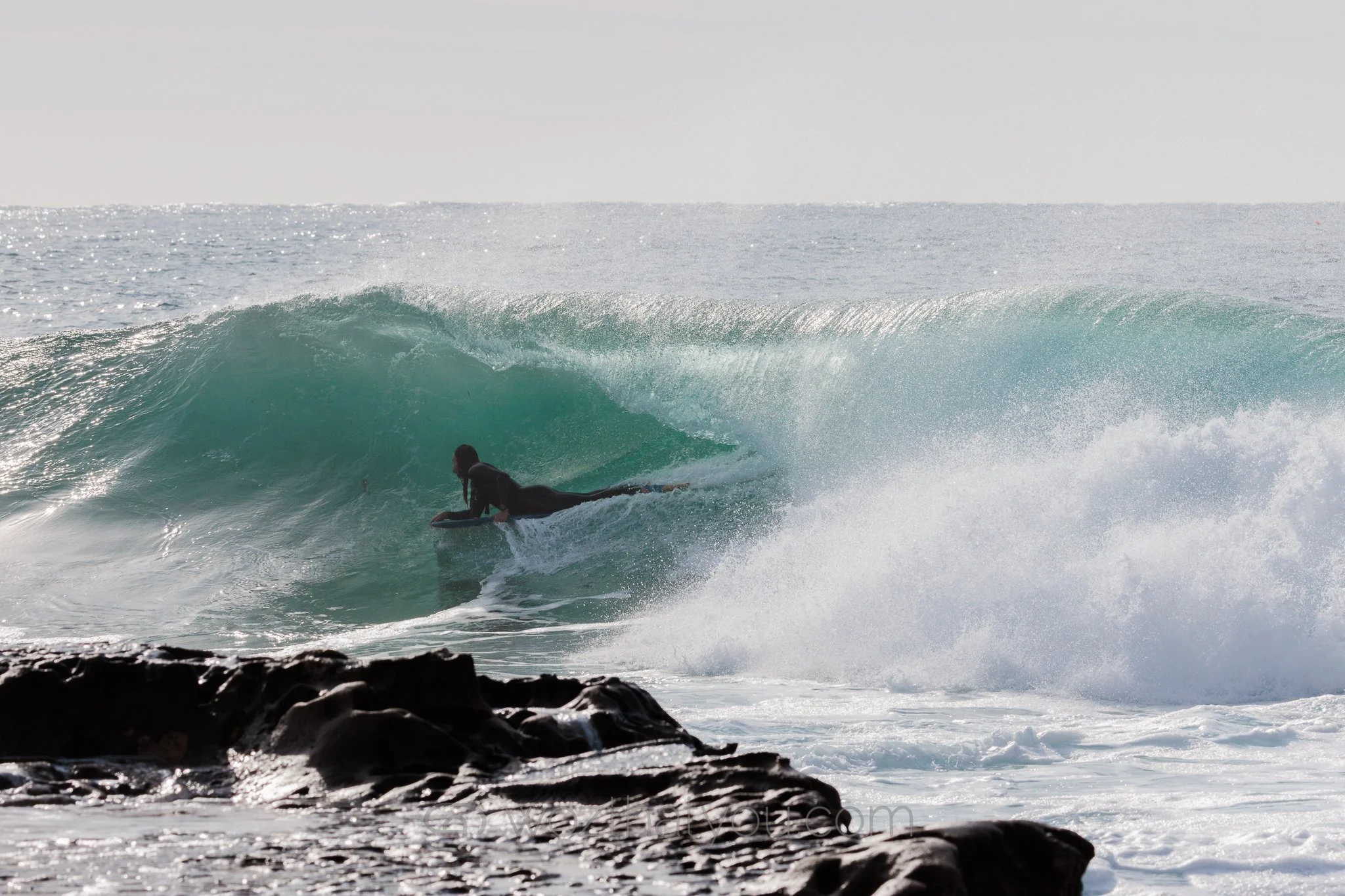 A surfer lying on a surfboard rides a wave near a rocky shoreline with the ocean in the background.