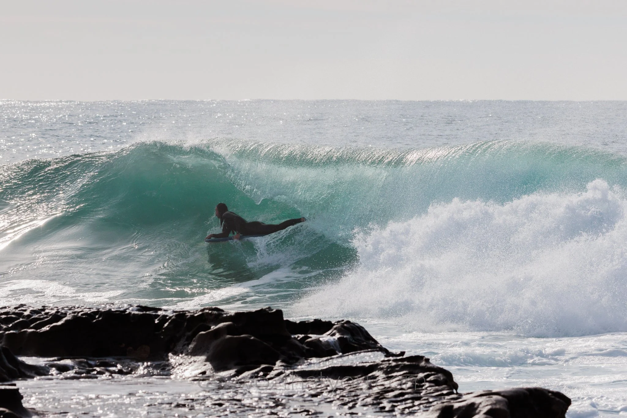 A person in a wetsuit surfing a wave near rocky shoreline with the ocean and horizon in the background.