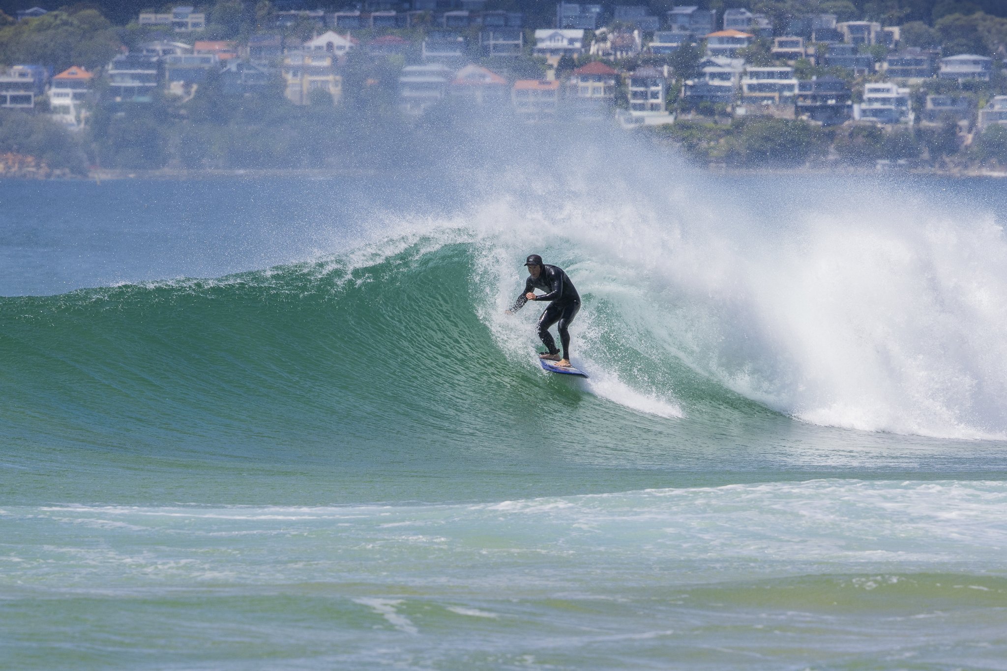 A person surfing on a wave near a shoreline with houses on a hill in the background.
