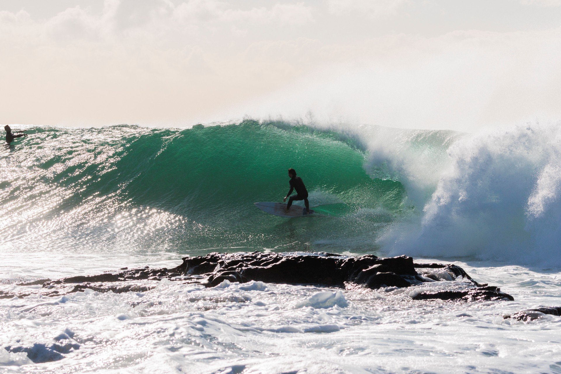 A person surfing on a large wave with another surfer on a surfboard in the background.