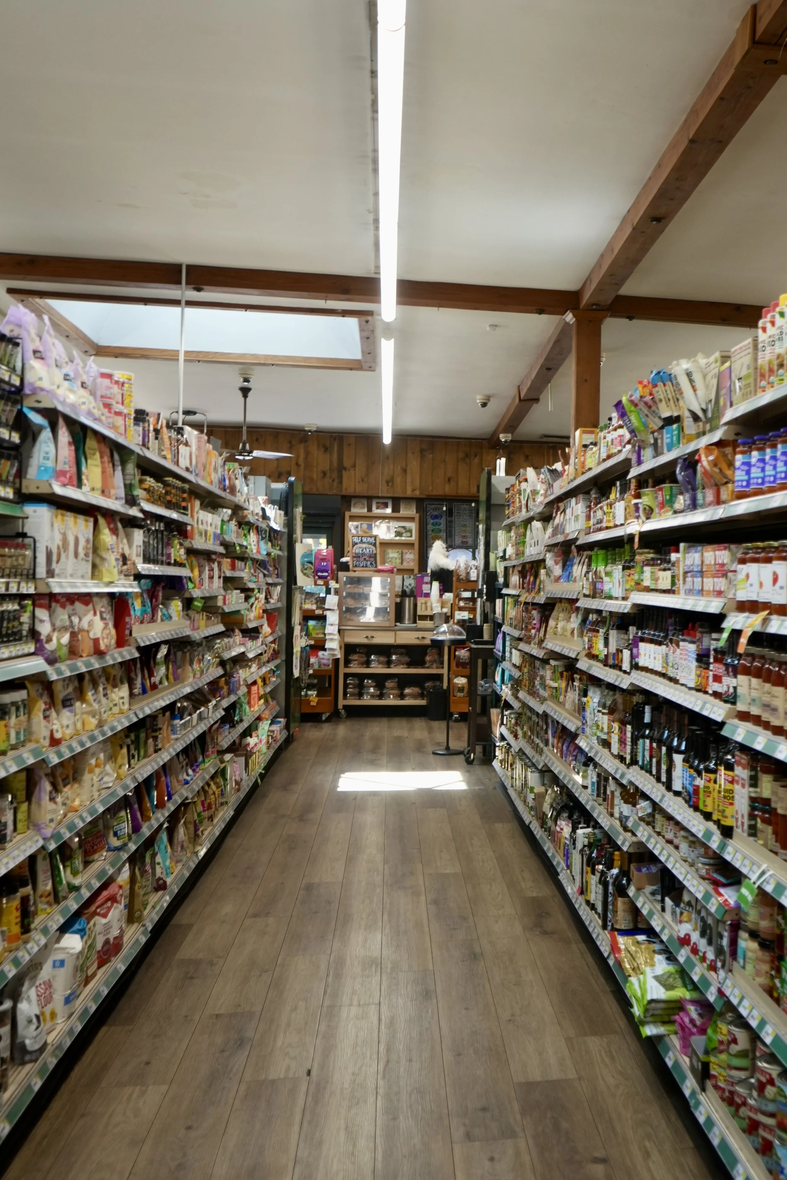 A green shopping basket containing various food items, including a wrapped turkey, containers of potato salad and another dish, a large bag of organic kelp snacks labeled '12 tides', a chocolate latte box, and a jar of what appears to be coffee or tea, on a tiled floor.