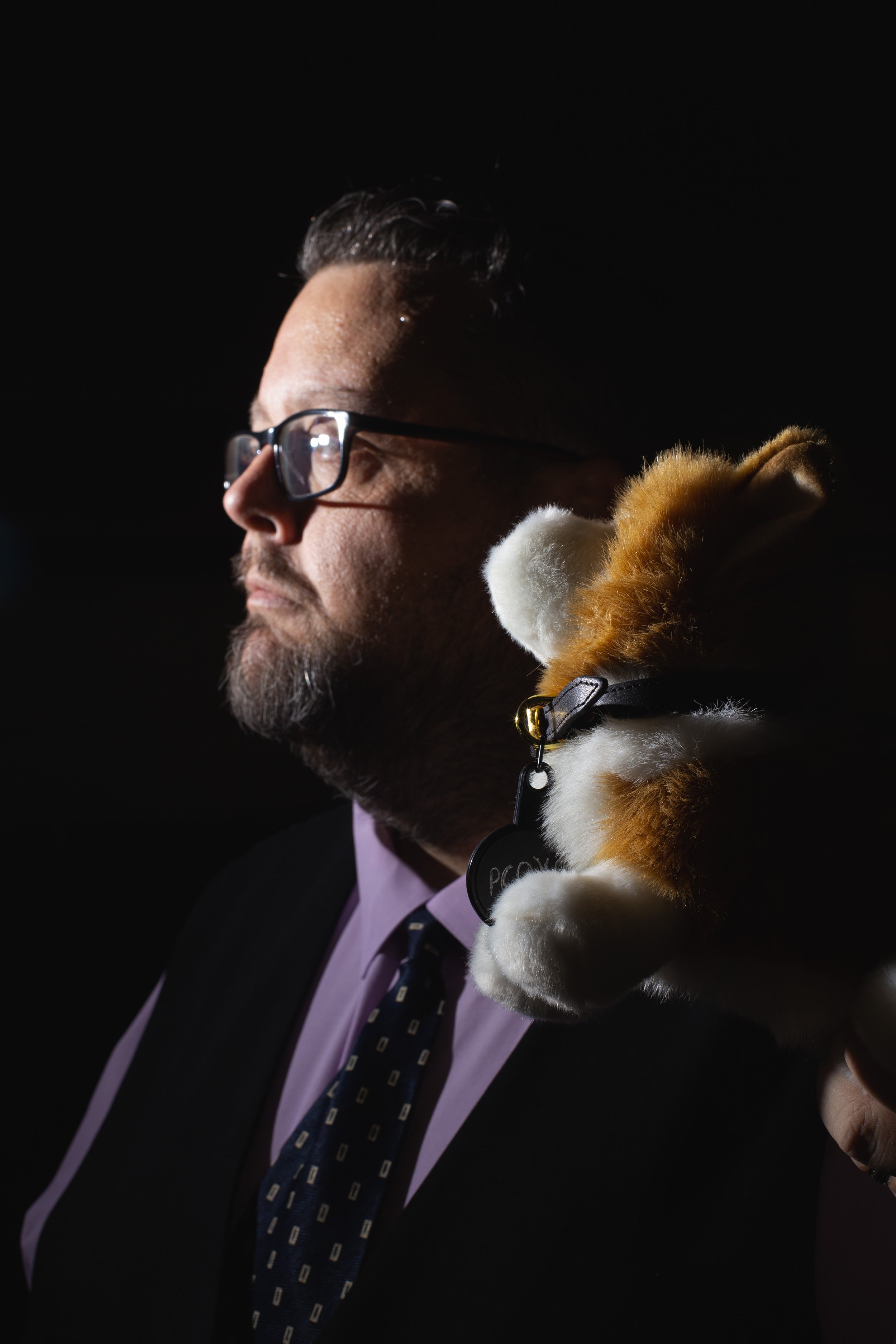 A man with glasses wearing a suit and tie sitting in a dark background with a plush toy dog on his shoulder.