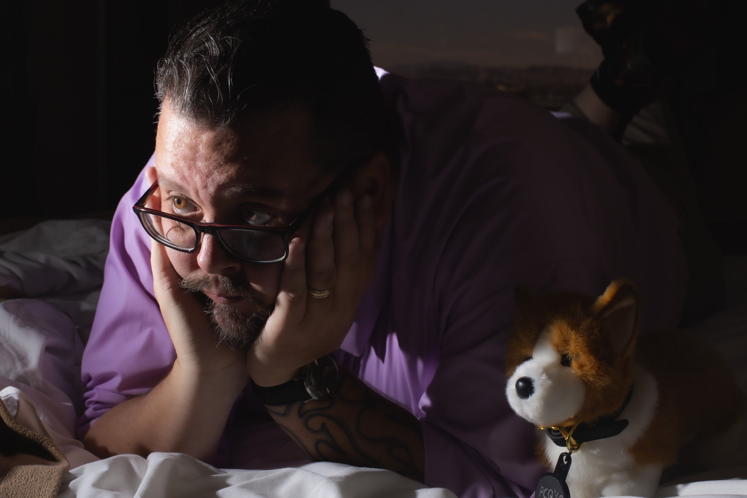 Marketing image of Jeff Walsh Comedy Magician lying on his stomach, resting his head on his hand, with a lion plush toy nearby in a dimly lit room.