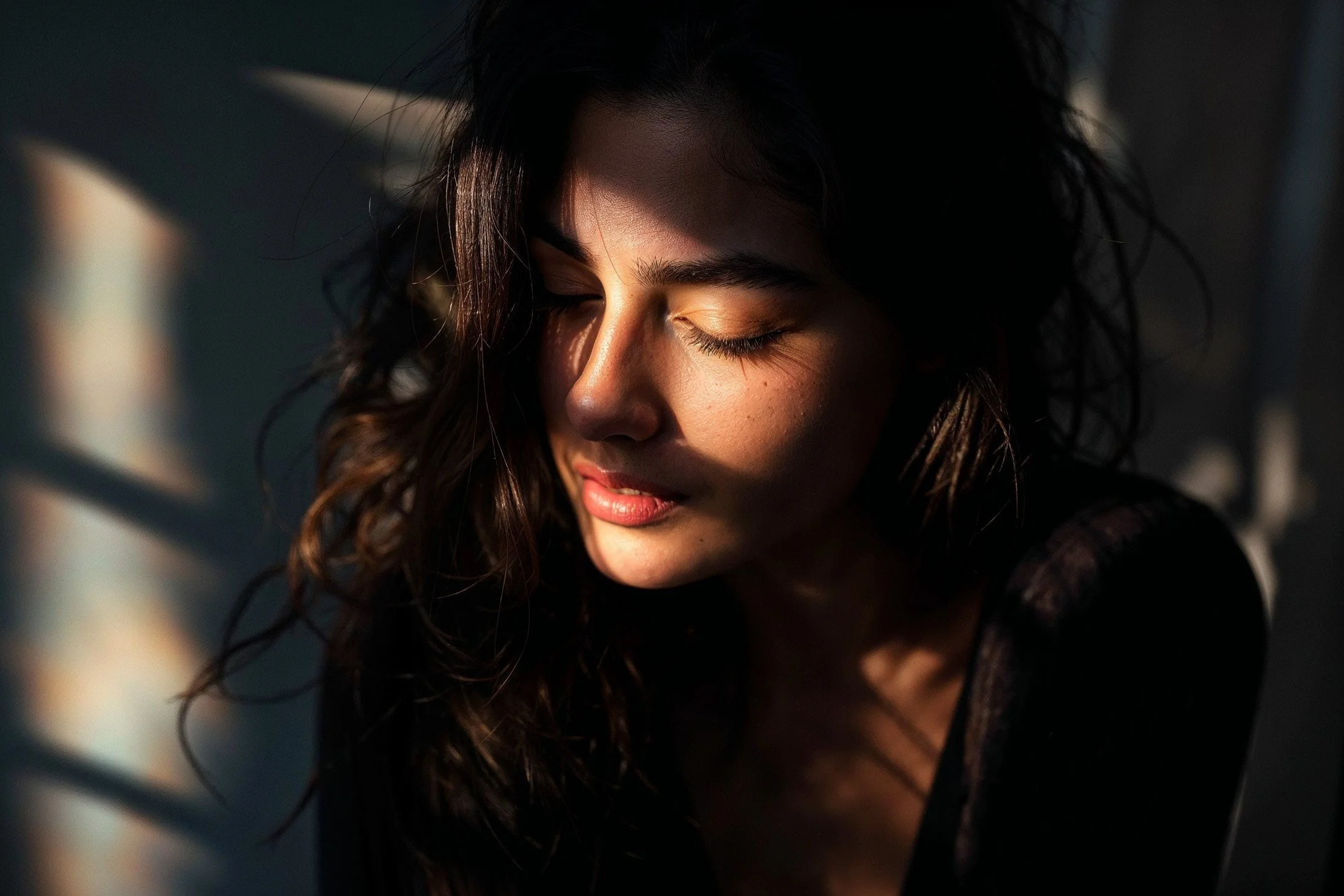 A woman with wavy dark hair and closed eyes, illuminated by soft light and shadows.