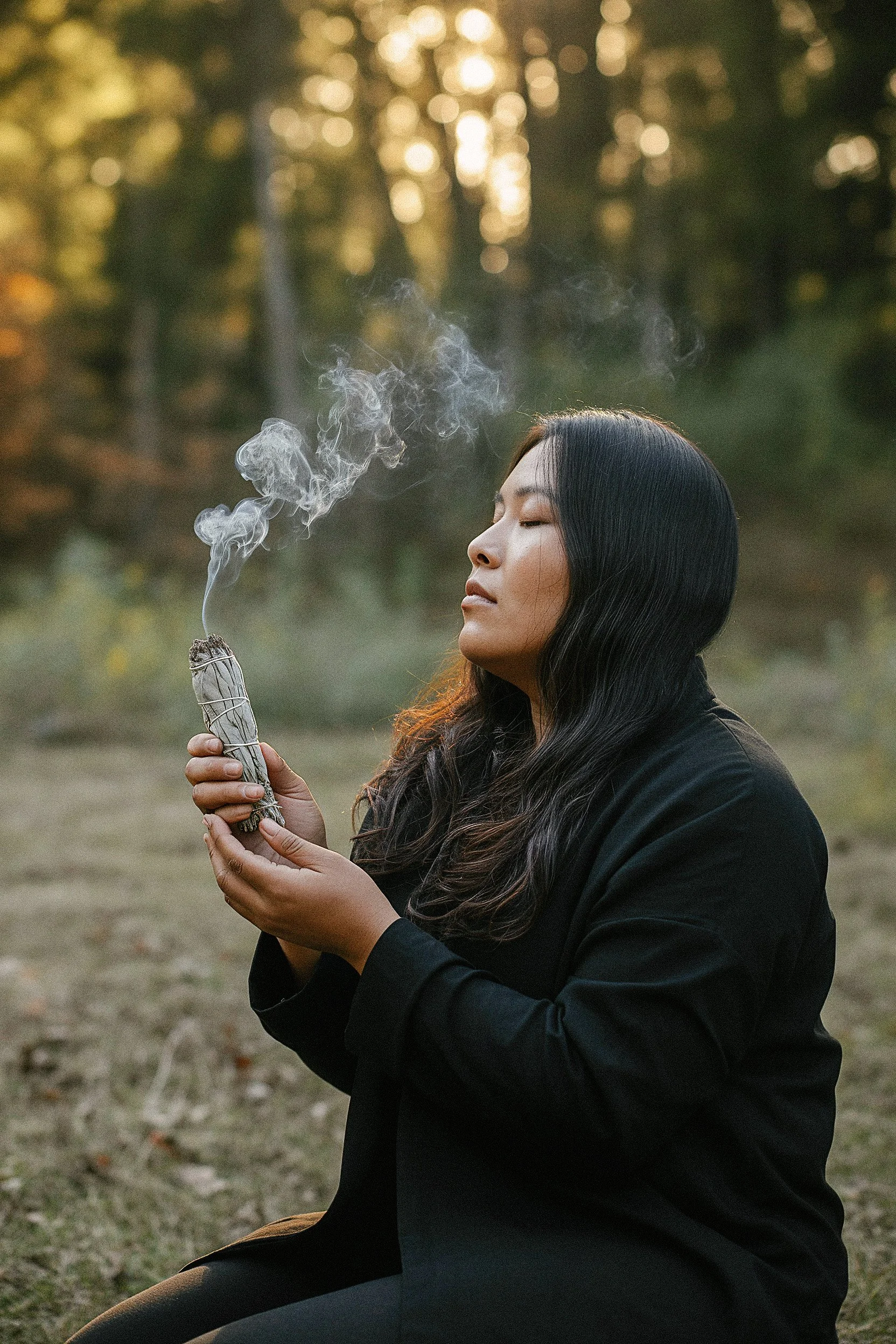 A woman with long dark hair and a black outfit is sitting outdoors in a natural setting during sunset or twilight, holding a smudging stick emitting smoke, with her eyes closed in a meditative or peaceful pose.