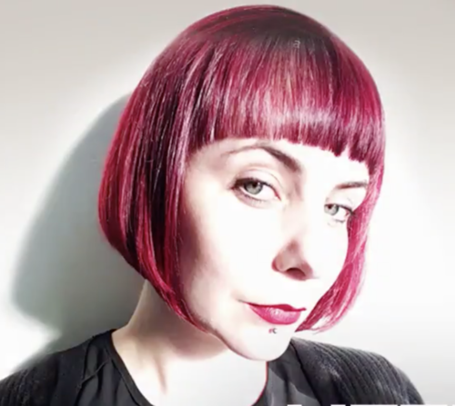 Woman with short, bright red hair and bangs, wearing a black top, looking at the camera with a slight smile.