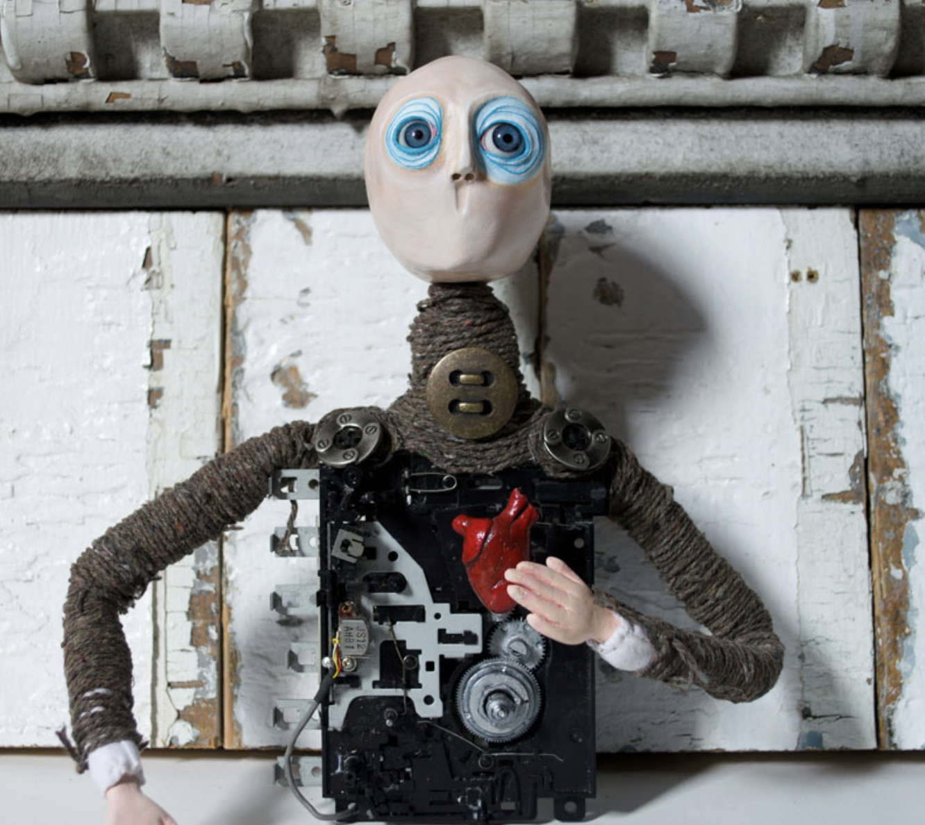 A humanoid robot with a human-like head, painted blue eyes, and a body made of mechanical parts and rope, holding a red heart in its hand, standing against a white, textured wall.