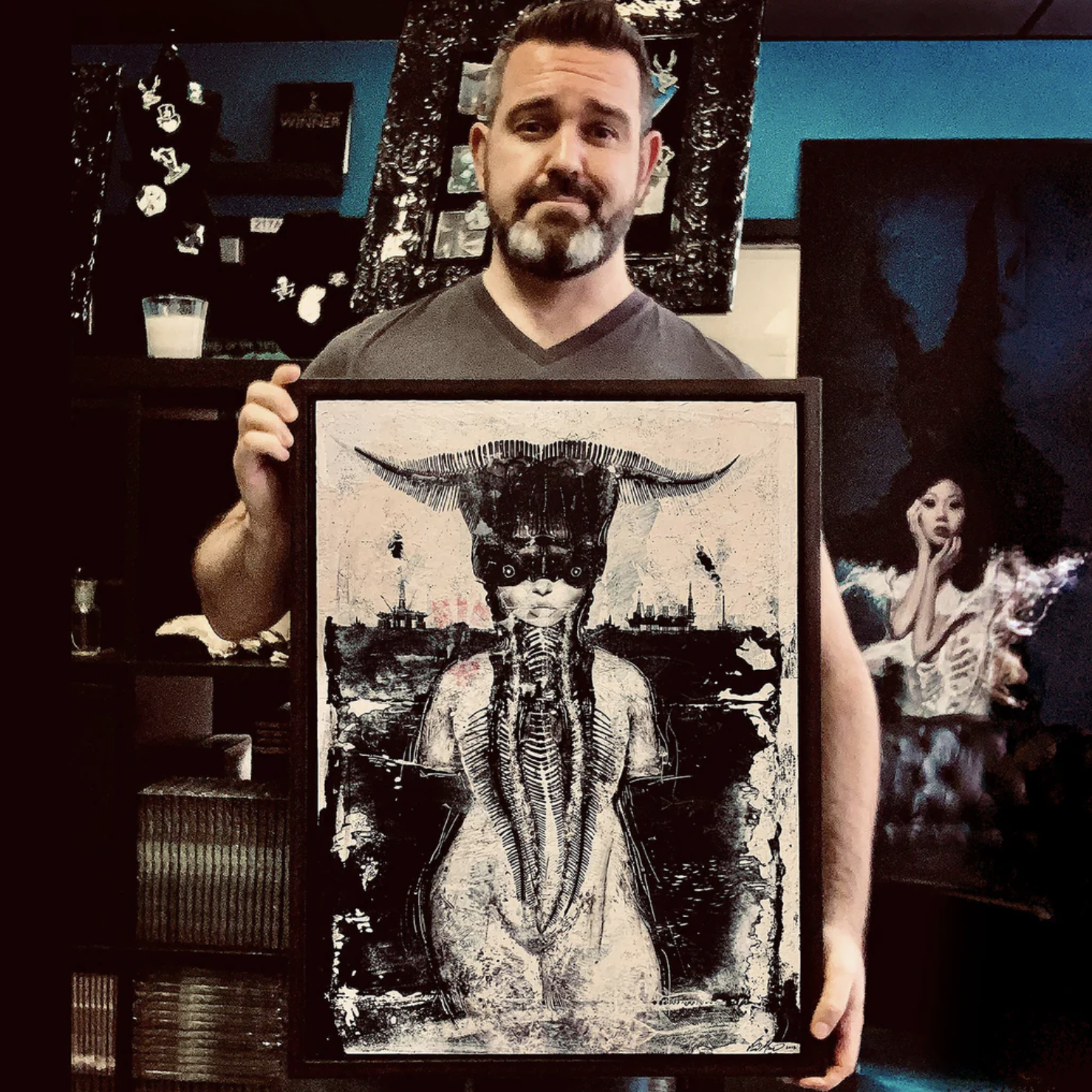 A man with a beard and mustache holding a framed artwork of a surreal figure with horns, a large eye on the face, and elongated features, in an art studio or gallery setting.