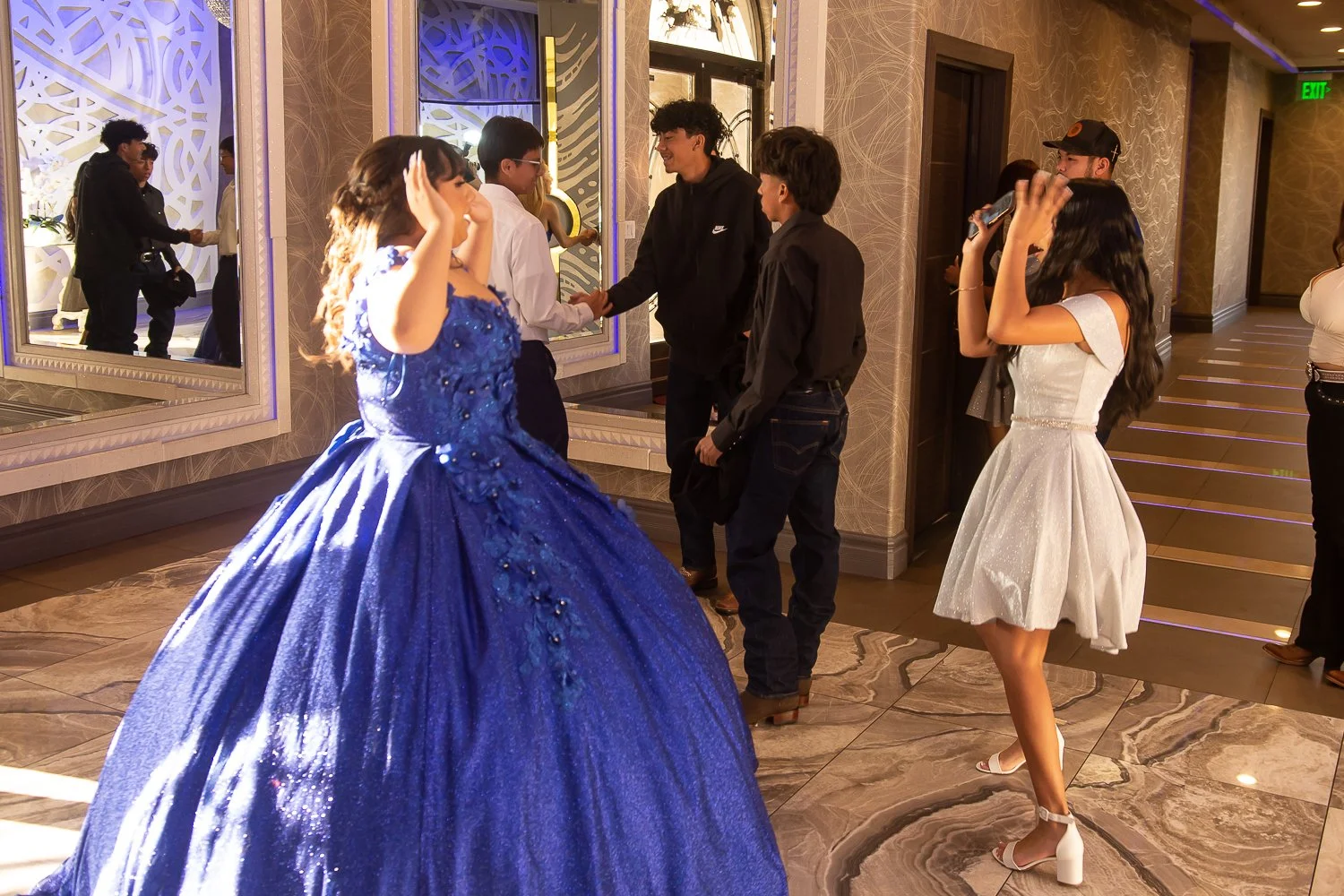 Event photography. Quinceañera. A young woman in a sparkling white dress takes photos of a girl in a blue ball gown at an event. Several other people are present, with two men shaking hands in the background as they look at each other.