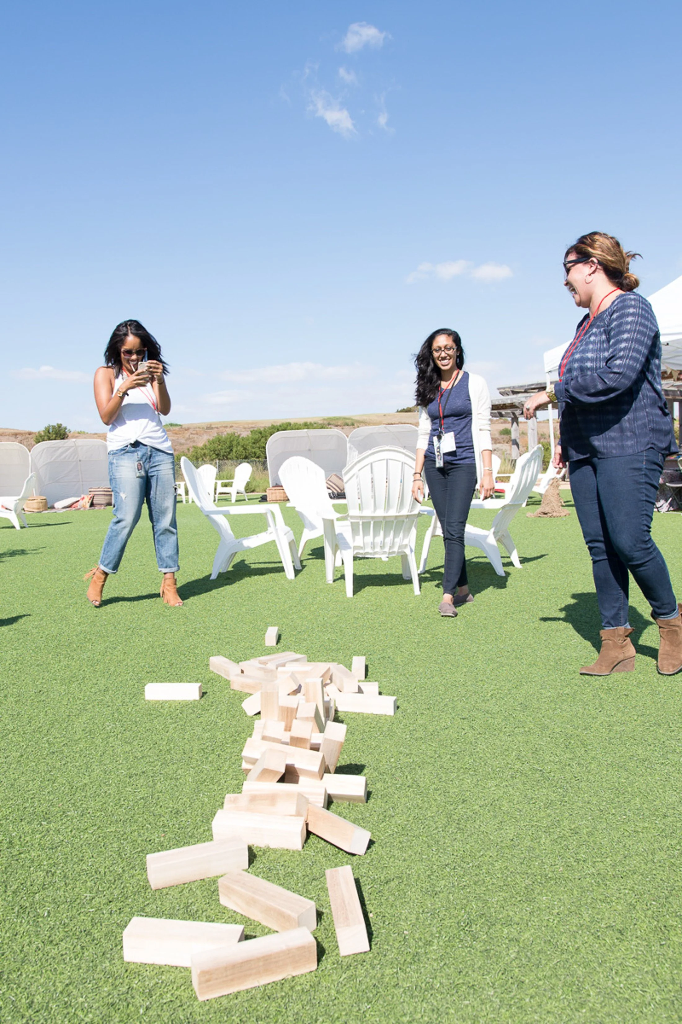 Three women are outdoors playing giant Jenga on green artificial grass and enjoying a sunny day. One woman is taking a photo, and two are smiling. There are white chairs and tables in the background under a clear blue sky.