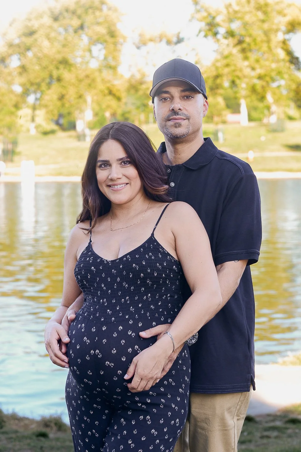 A pregnant woman wearing a navy blue dress with white floral patterns, smiling, stands in front of a man, both embracing near a body of water with trees and a park in the background.