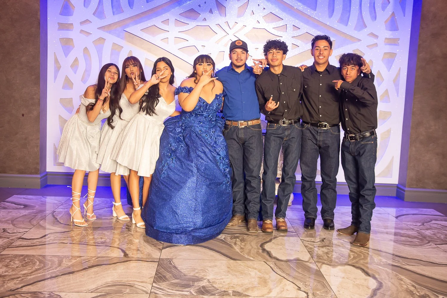 Event photography. Quinceañera. A group of eight young people dressed in formal attire posing together in front of a decorative backdrop. Four women in white and a blue gown and four men in black and blue shirts with jeans.