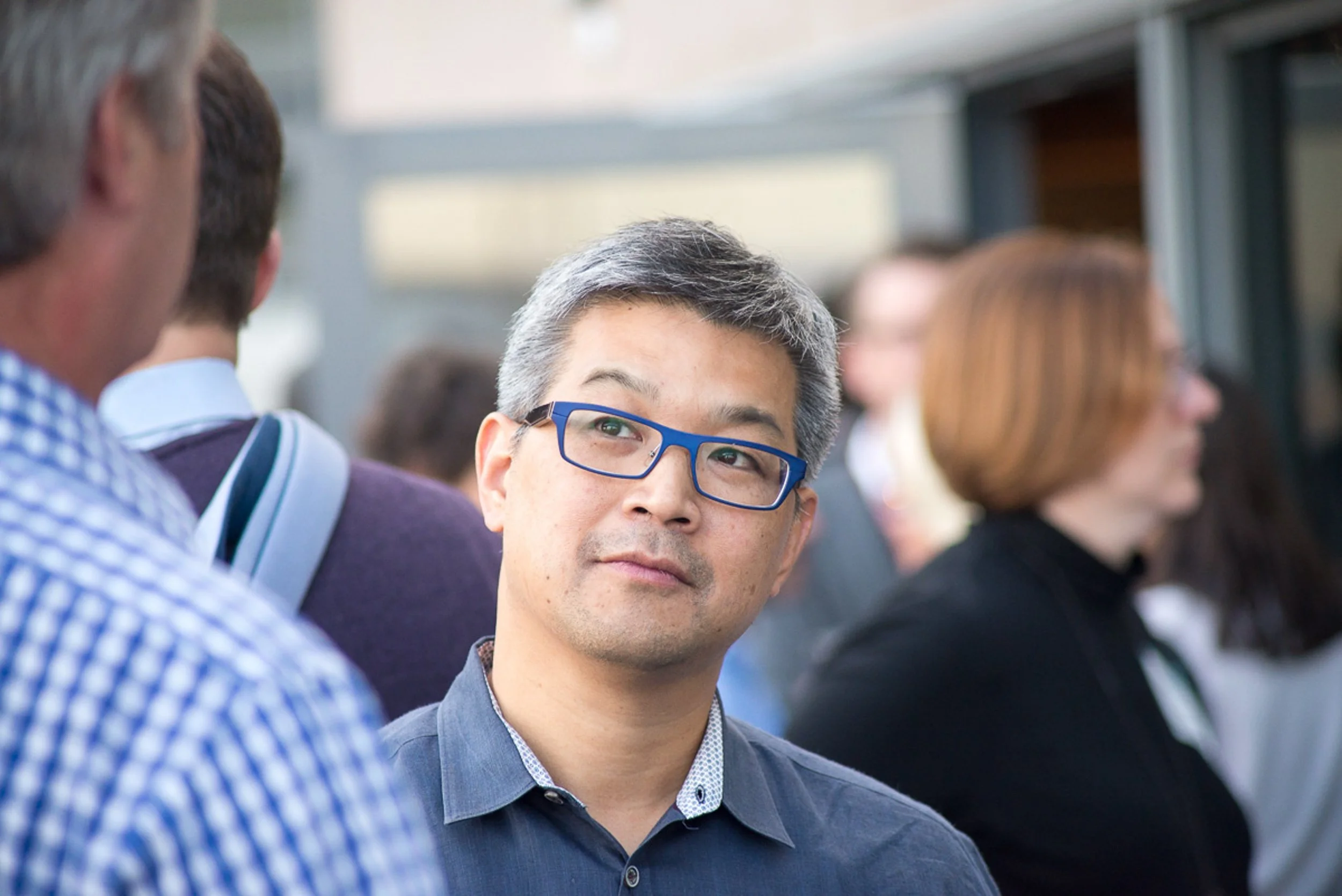 Event photography. A man with gray hair, glasses, and a dark shirt, standing among a group of people, looking attentively to his left, in an outdoor setting.