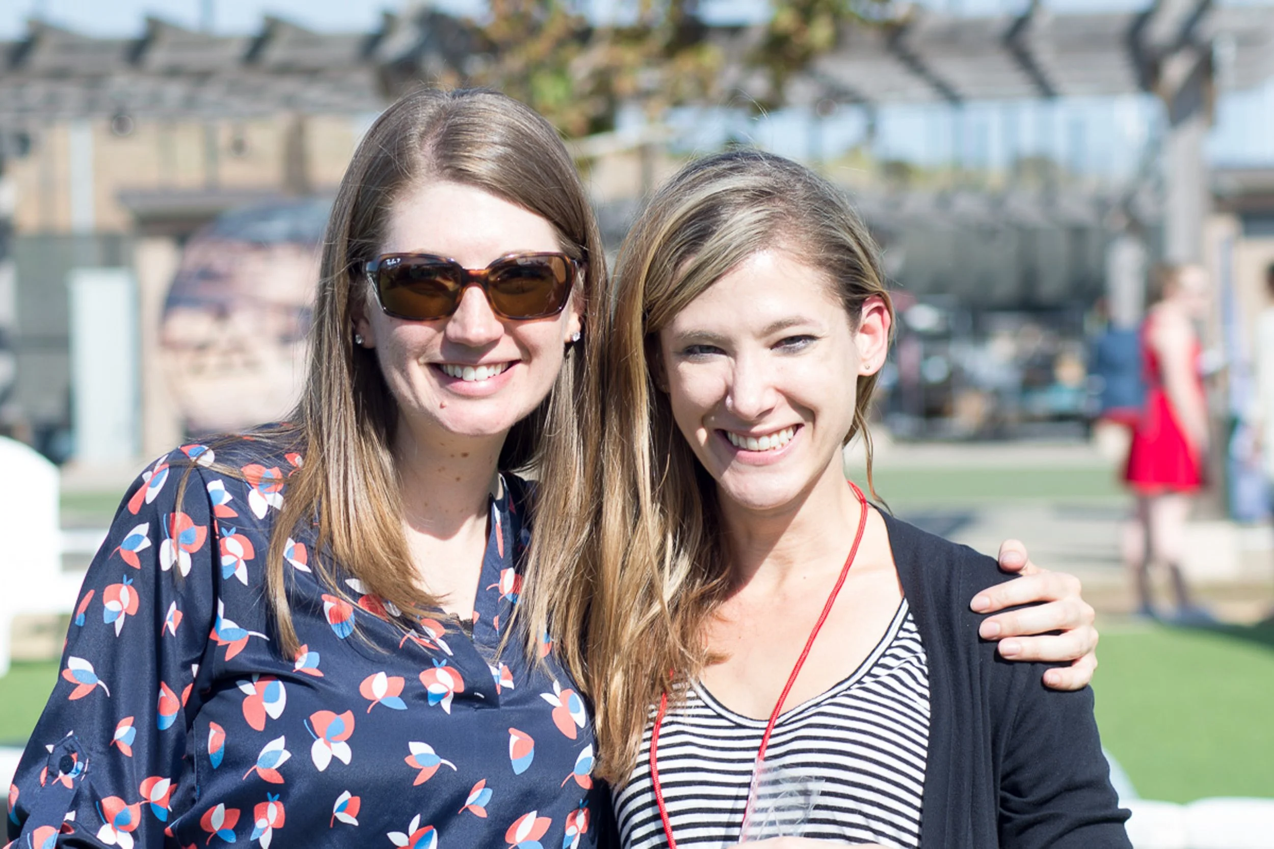 Two women smiling outdoors, one wearing sunglasses and a patterned shirt, the other wearing a black cardigan and striped top, with a blurred background of people and structures.