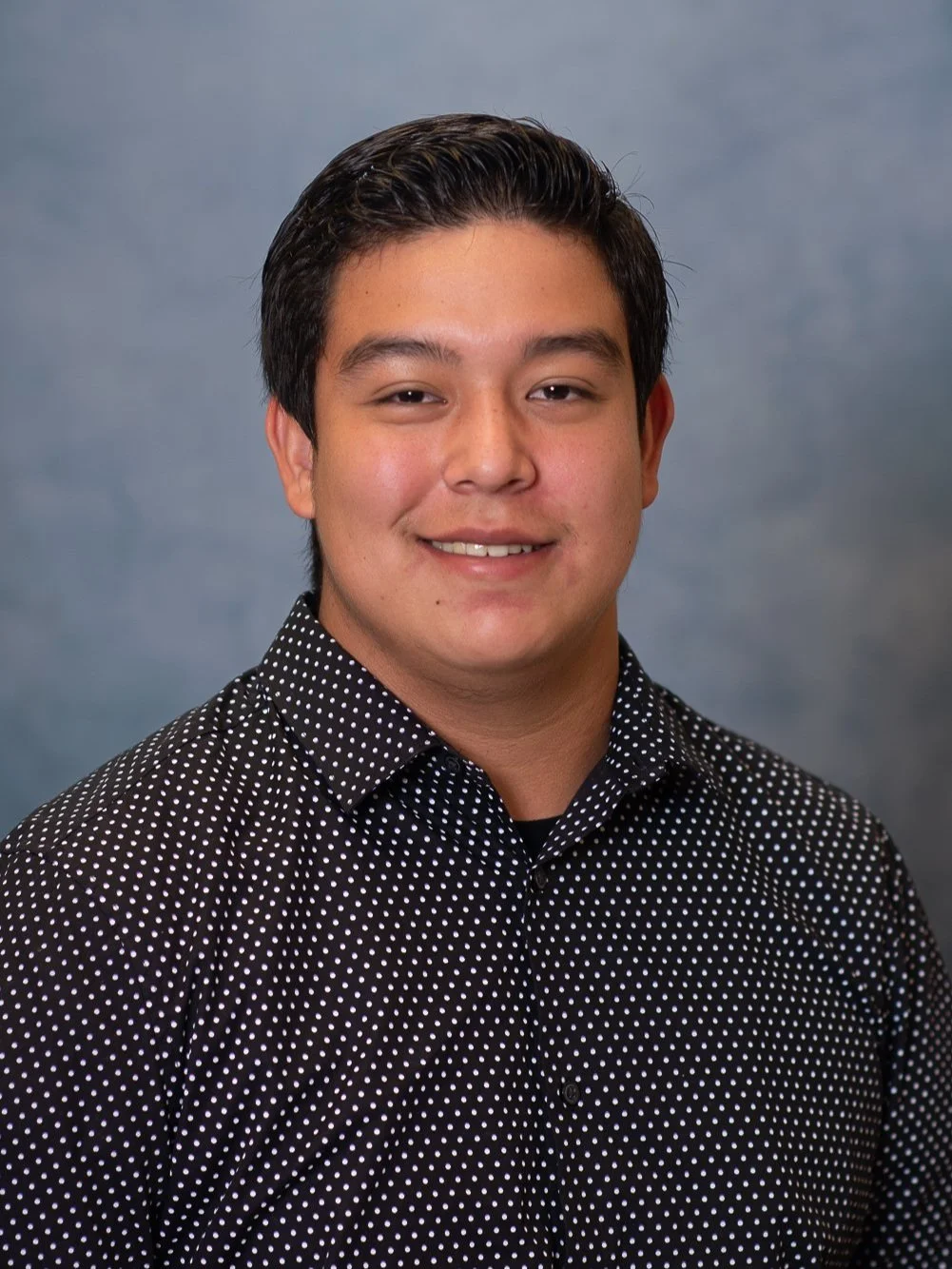 School photography. Portrait of a young man with dark hair, smiling, wearing a black collared shirt with white polka dots, against a gray background.