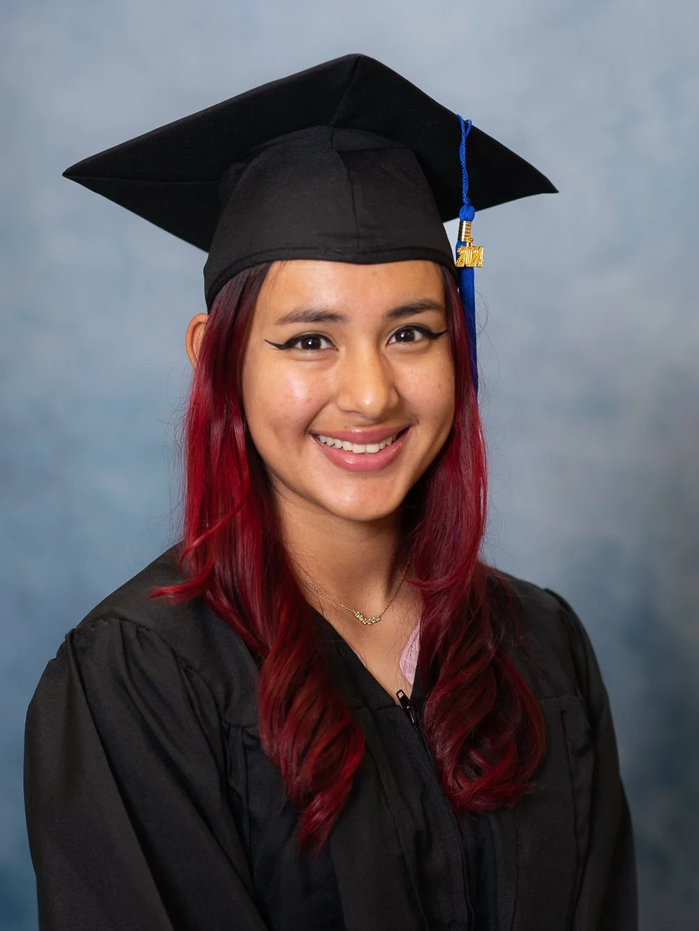 Cap and gown photography. A young woman with red hair smiling, wearing a graduation cap and gown, against a blue background.