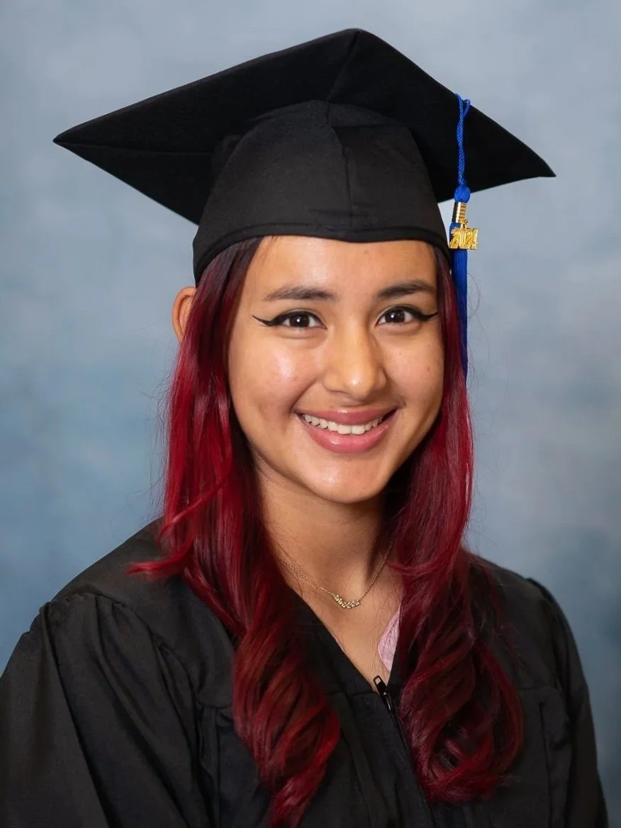 Cap and gown photography. A young woman with red hair smiling, wearing a graduation cap and gown, against a blue background.