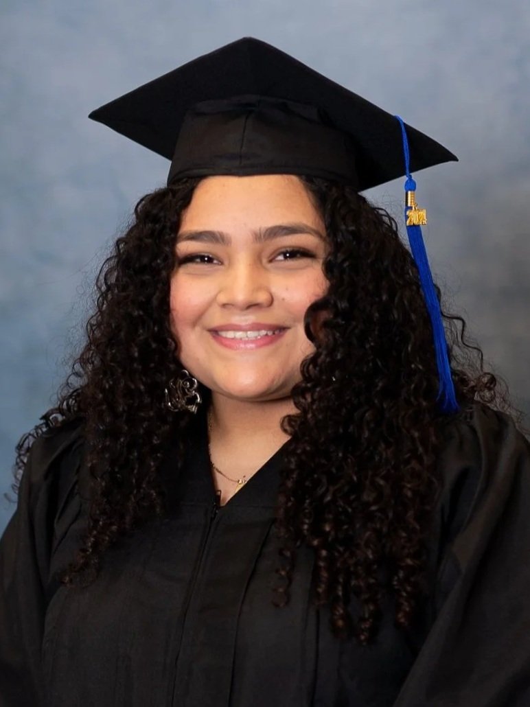 Cap and gown photography. Young woman smiling in graduation cap and gown against a blue background.