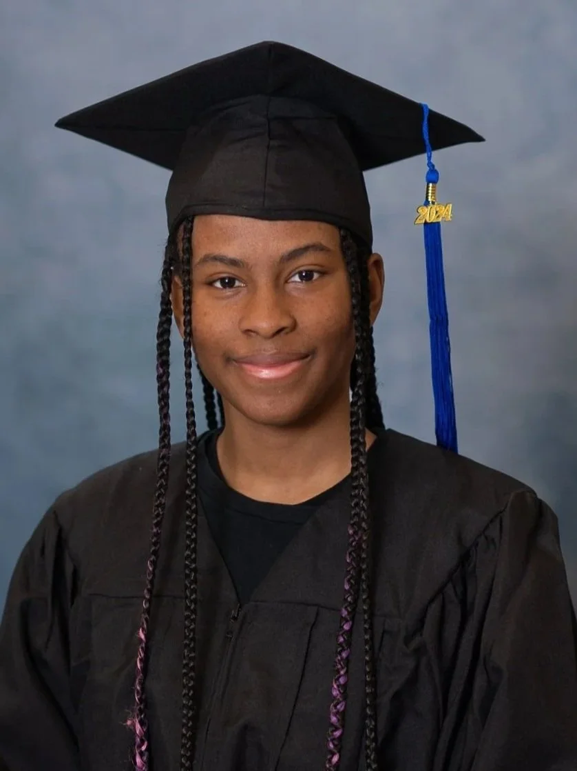 Cap and gown photography. Young woman in black graduation gown and cap, with blue tassel and a gold '2024' charm, smiling against a blurred blue background.