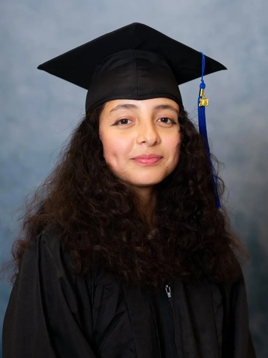 Cap and gown photography. Young woman in a black graduation cap and gown, standing against a gray background.