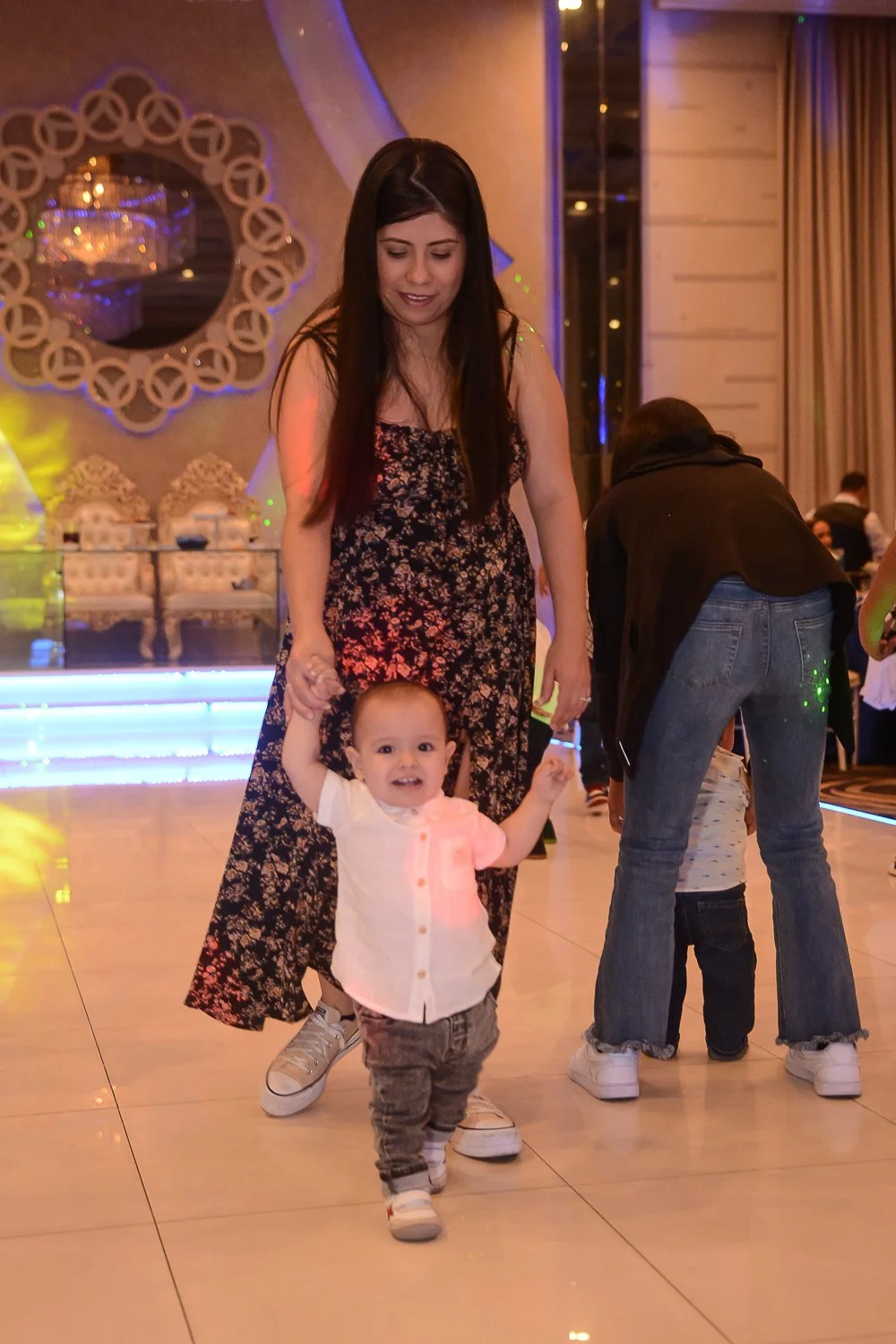 A woman and a young boy are together in a decorated indoor event space. The woman is holding the boy's hand, and the boy appears happy, smiling at the camera. The background features elegant decor and ambient lighting.