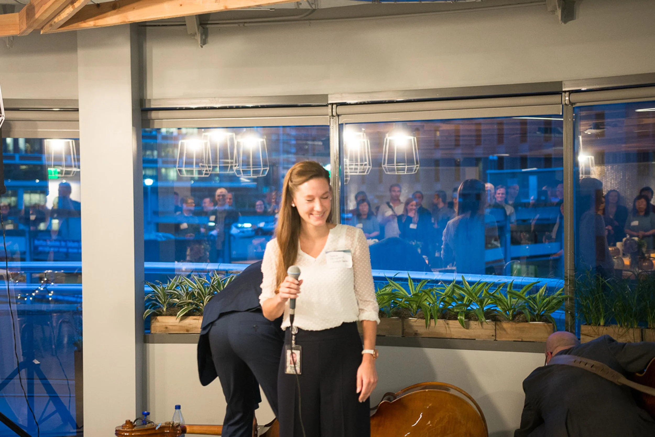 A woman holding a microphone smiling in an indoor event with a large window revealing a crowd outside.