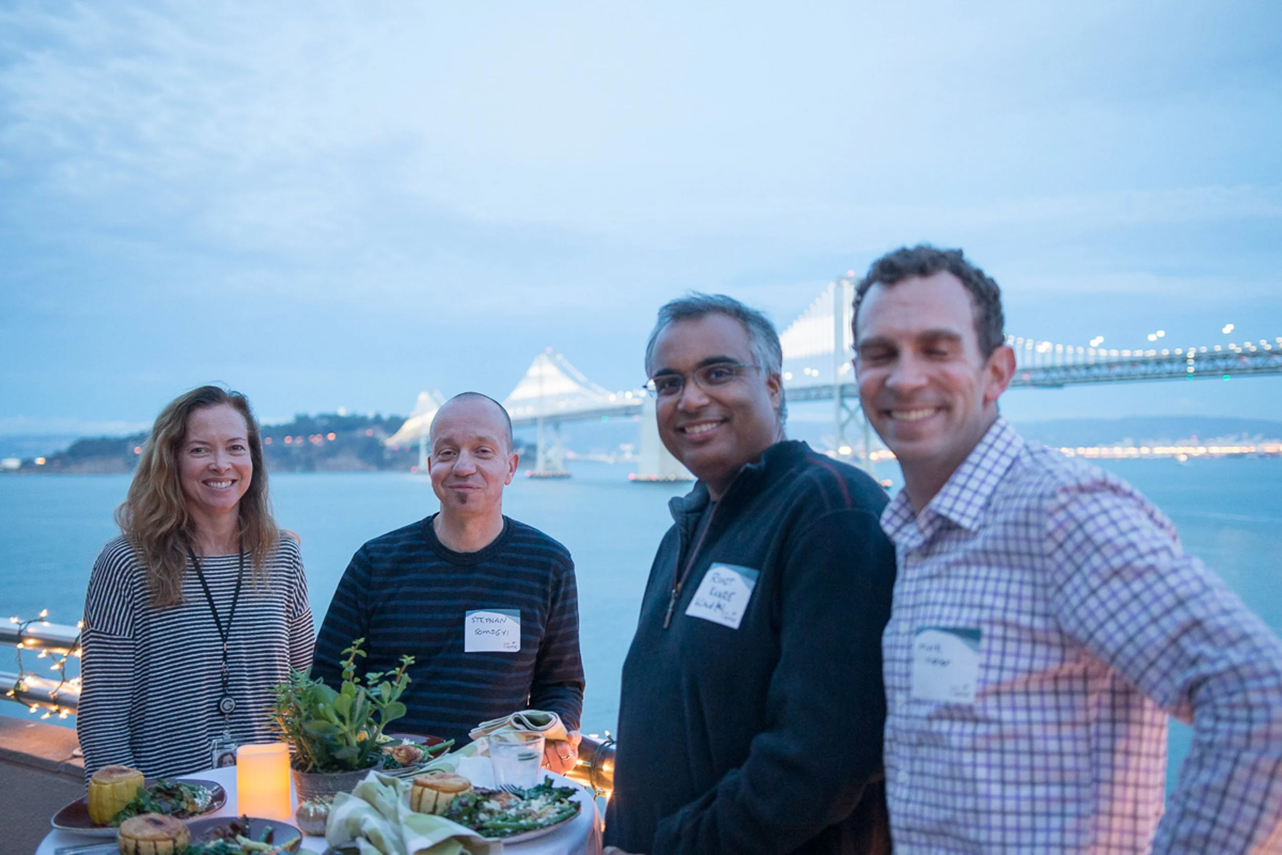 Four people standing outdoors near water with a bridge in the background, smiling, with a table of food and decorations in front of them.