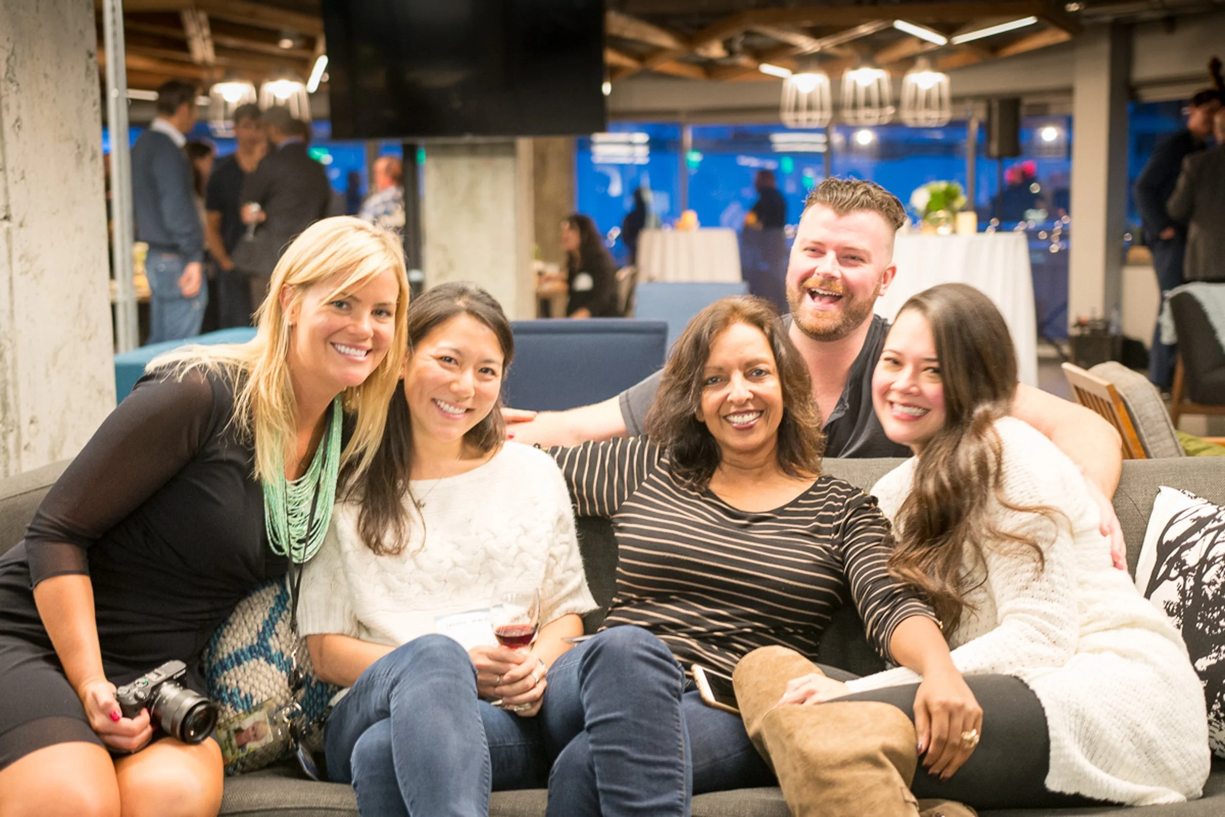 Group of five people sitting on a sofa at a social gathering, smiling and posing for the photo, with a bar or restaurant setting in the background.