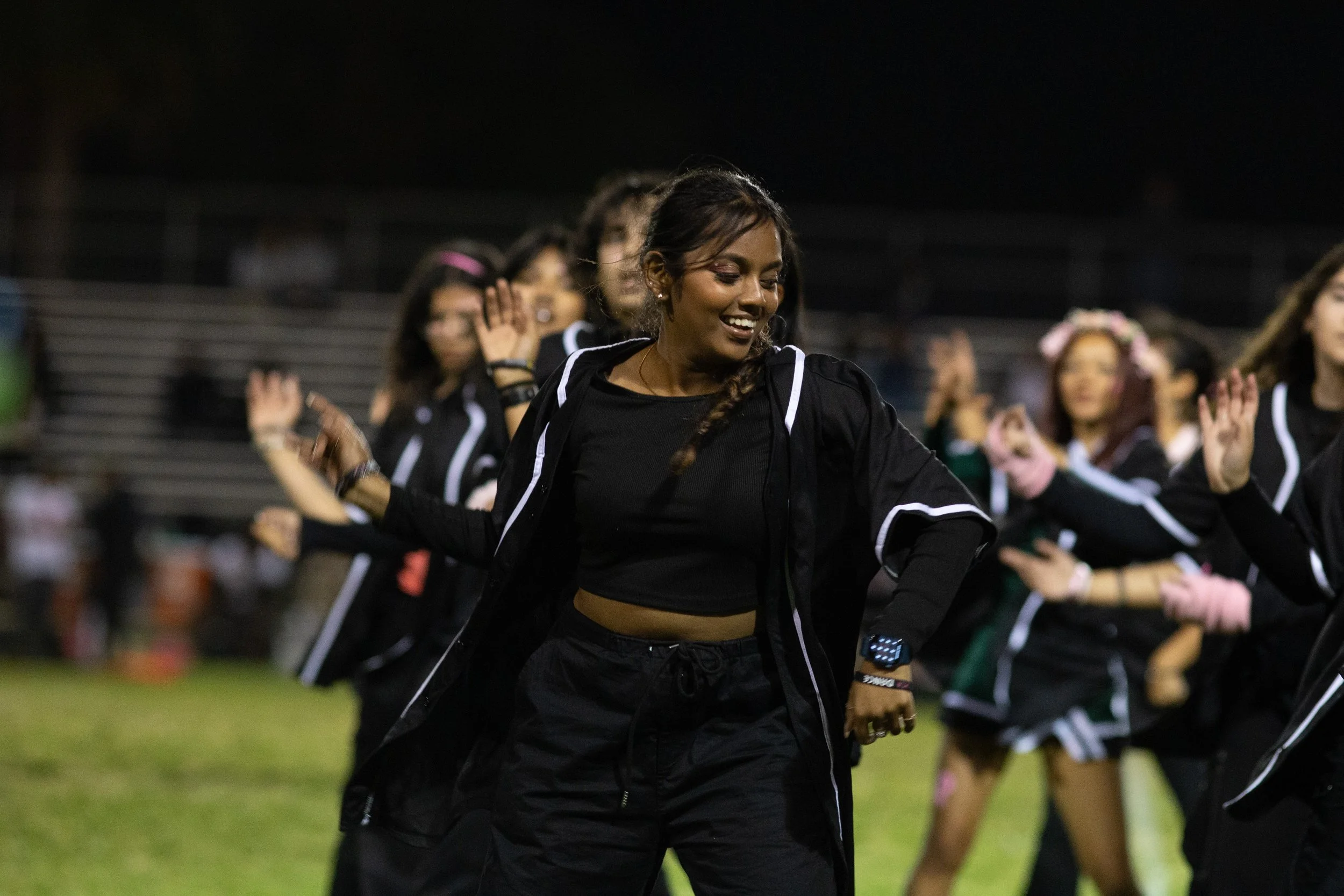 A group of women dancing on a field at night, wearing black sports jackets with white stripes, with one woman in the foreground smiling and enjoying the dance.