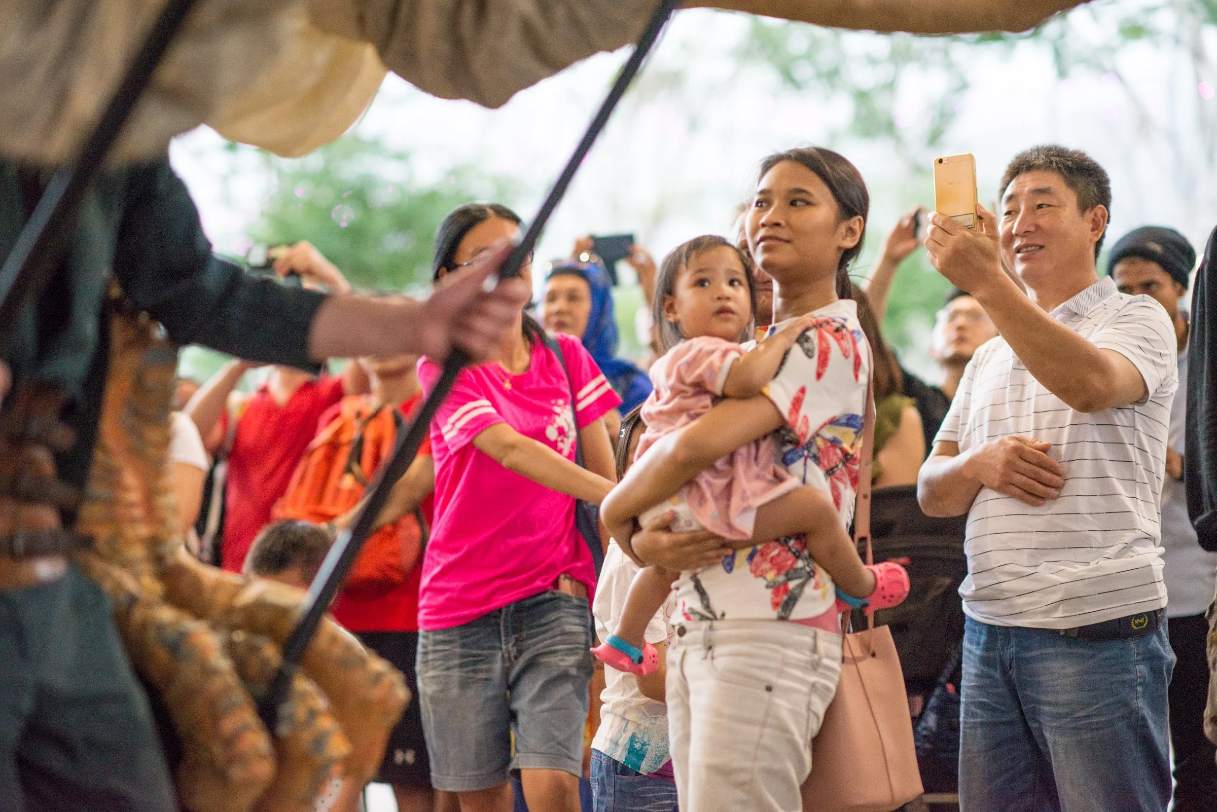 A woman holding a young girl, surrounded by people taking photos or videos with their phones, in an outdoor setting with trees.