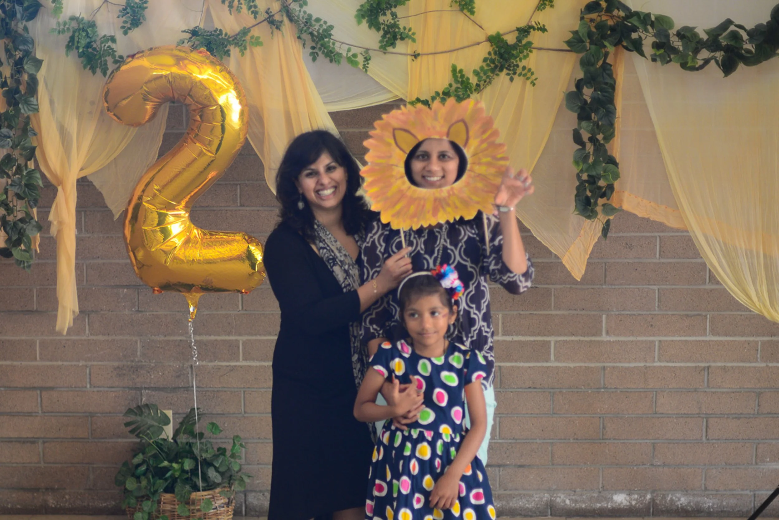 Event photography. A woman and two children celebrating a second birthday with balloons, decorations, and props, including a gold numbered balloon '2' and a face cutout of a lion.