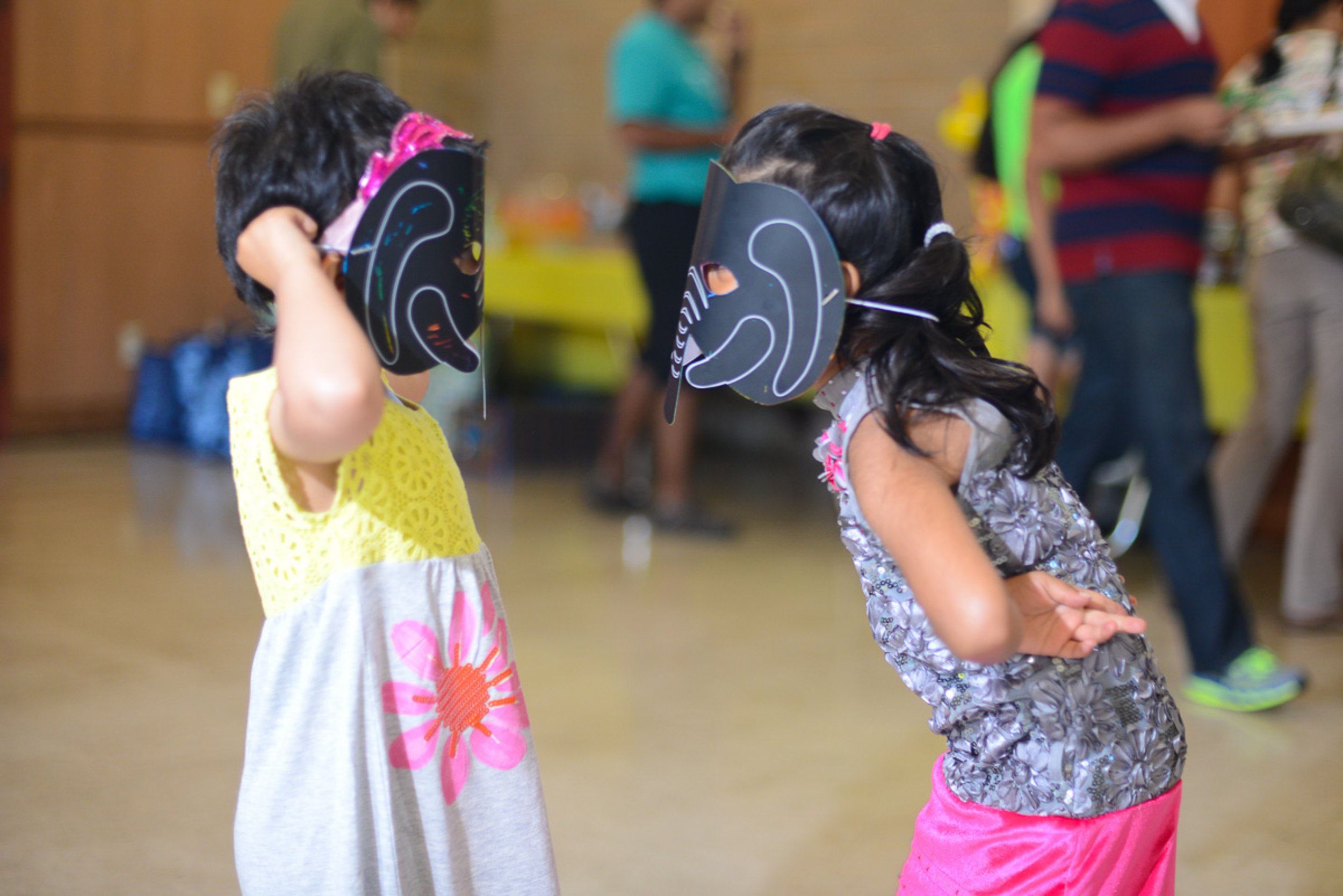 Two young children face each other wearing black masks of human ears, engaging in a playful activity at an indoor event.