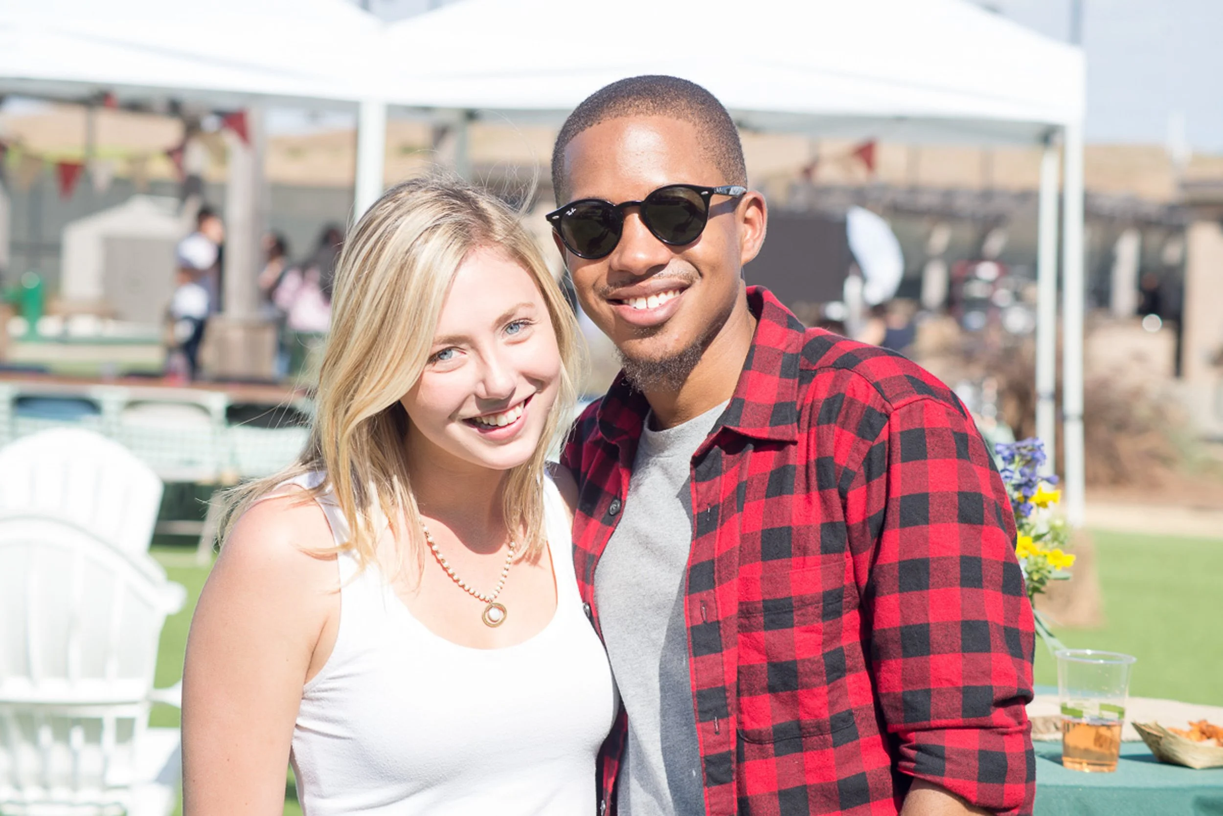 A smiling young woman with blonde hair and a necklace stands next to a smiling young man wearing sunglasses and a red plaid shirt, outdoors at a daytime event.