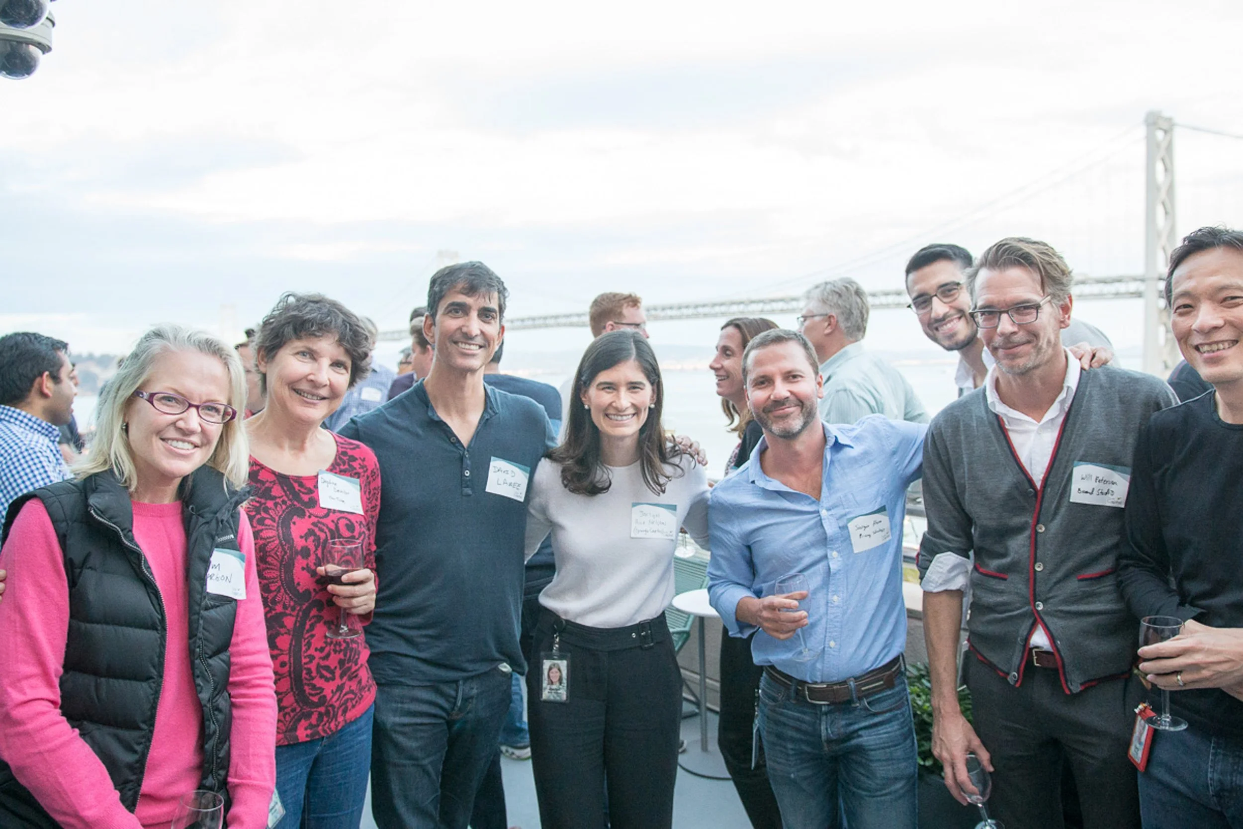 Event photography. Group of smiling people at an outdoor event, with a bridge in the background, holding glasses of wine.