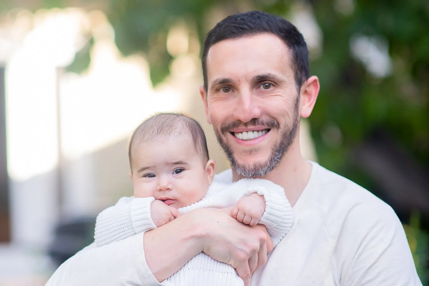 A smiling man with a beard holding a baby with a contemplative expression outdoors.