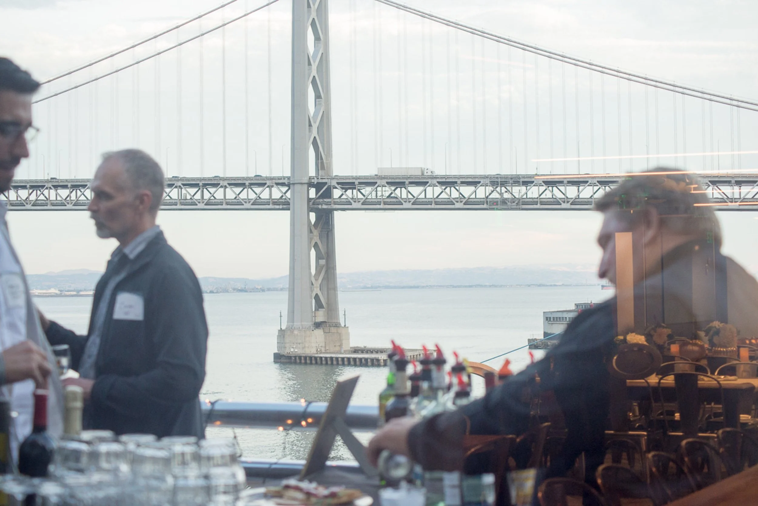 Event photography. View through restaurant window of the San Francisco-Oakland Bay Bridge with guests and restaurant setup inside.