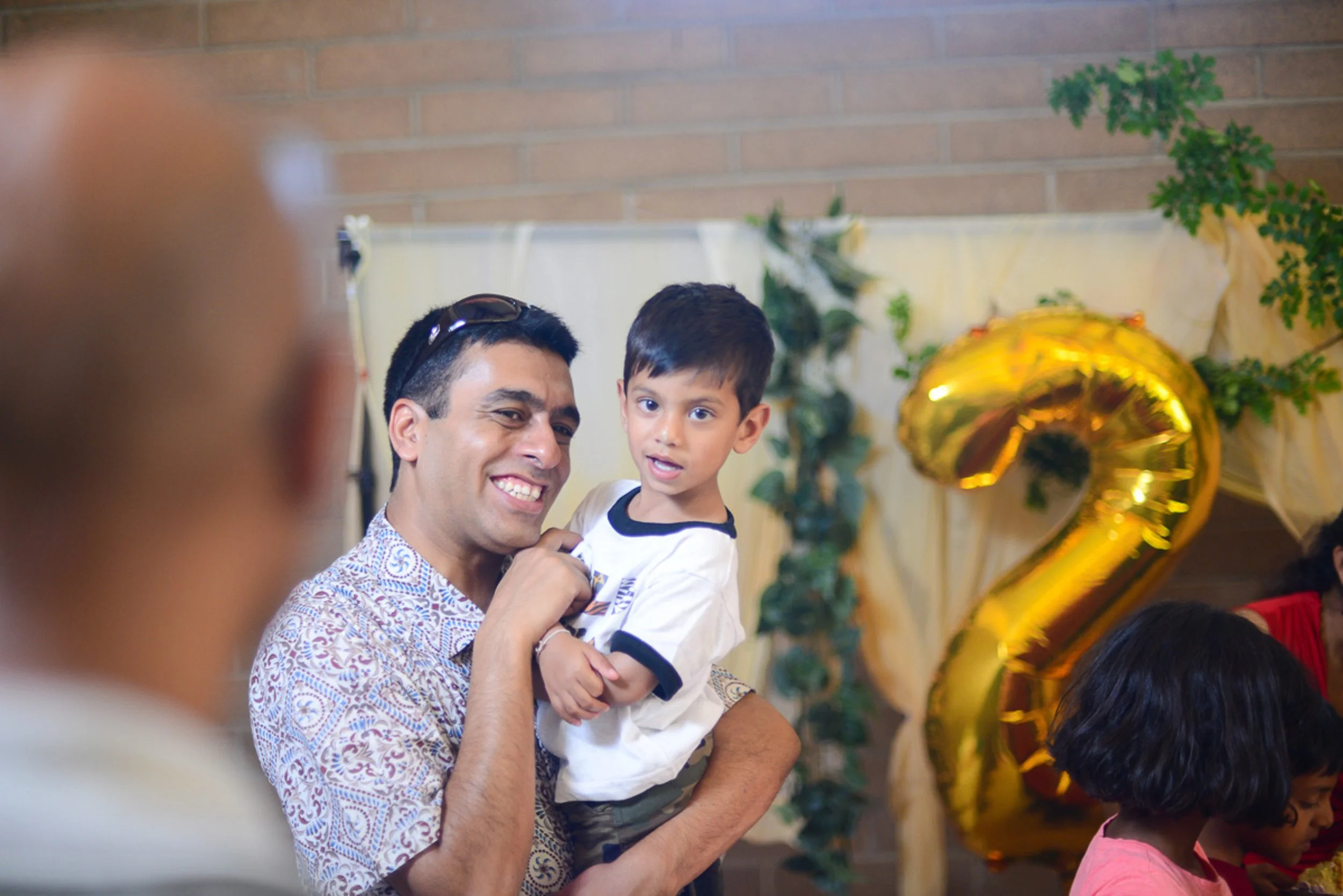 Event photography. A man smiling and holding a young boy at a celebration with large gold balloon shaped as number 2, greenery and decorations in the background.