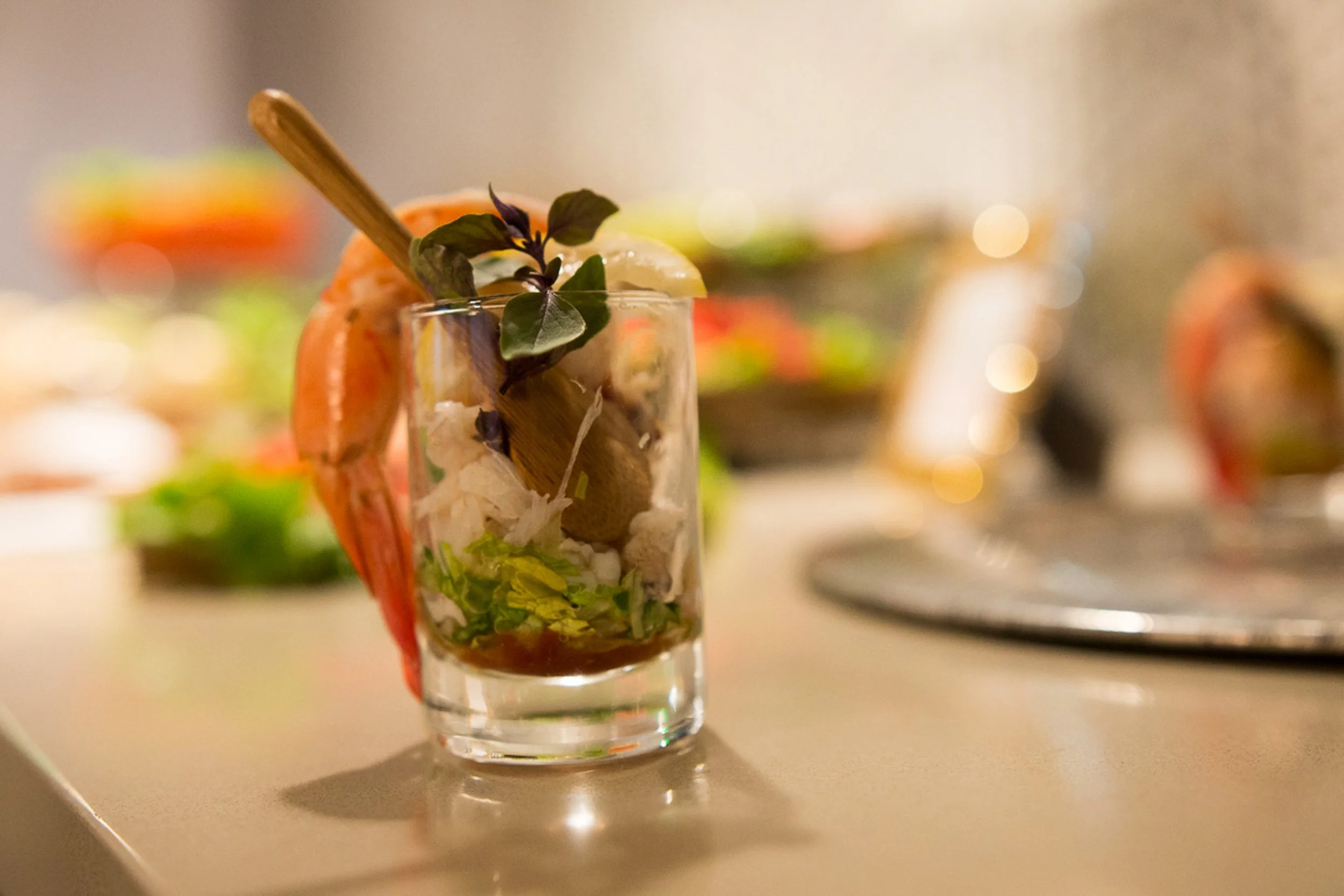Event photography. A glass with seafood, greens, and herbs, garnished with a sprig of basil and a lemon wedge, on a light-colored surface with blurred trays of food in the background.