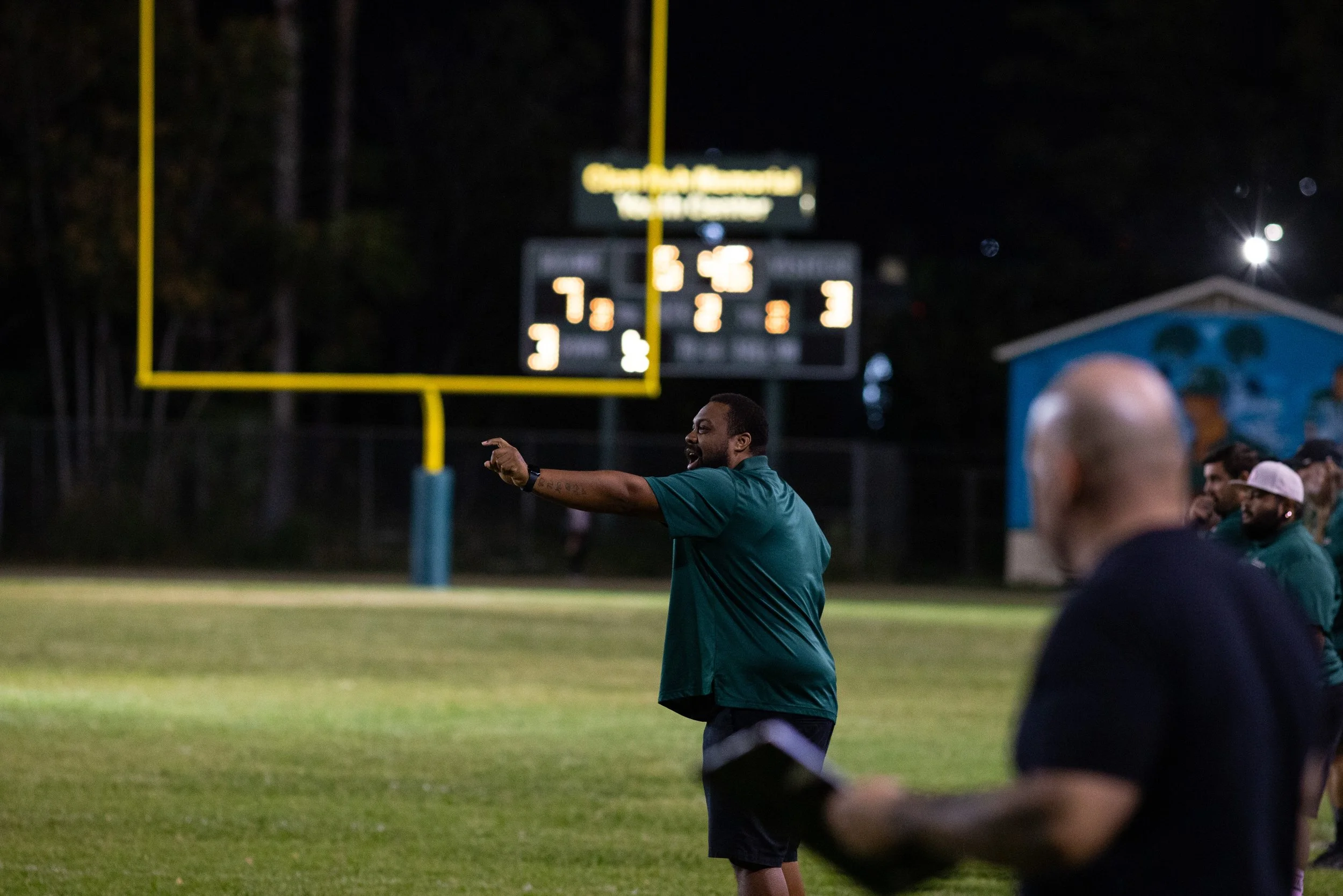 School photography. A football coach on the sideline of a football field during a game at night, gesturing and talking to players or officials. A large scoreboard and goalpost are visible in the background.