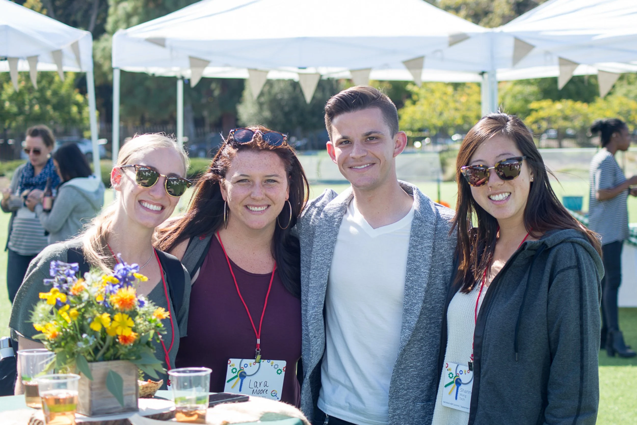 Four people standing outdoors at a sunny event, smiling for the camera. Two women on either side wear sunglasses, one woman wears a name tag that reads 'Lara Moore,' and a man in a gray jacket is between them. There are tents, other people, and trees