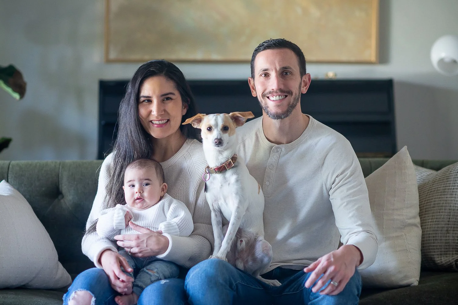 A happy family sitting on a green couch with a baby, a dog, and a woman, all smiling at the camera in a cozy living room.