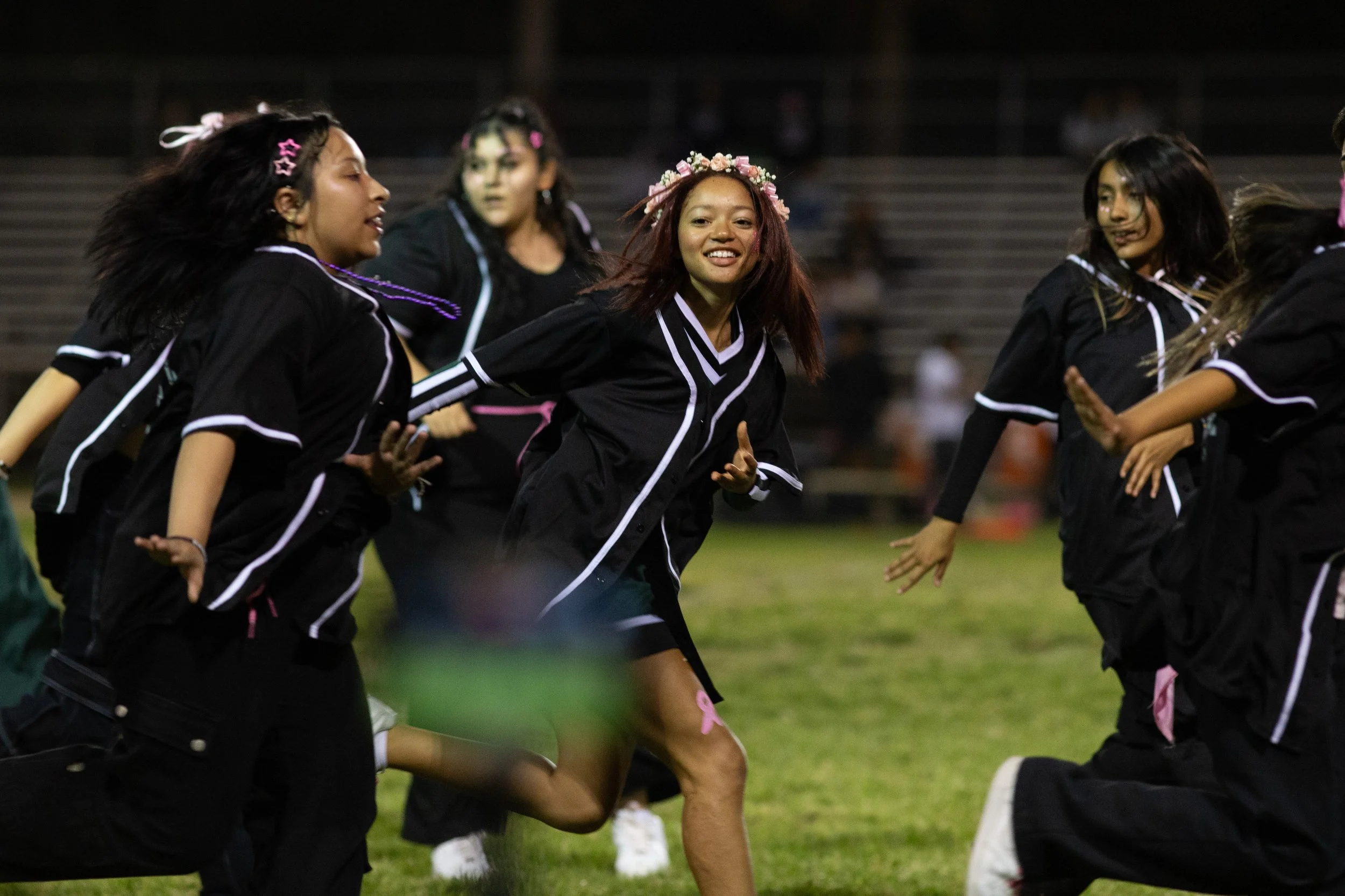 A group of young women wearing black jackets with white trim, participating in a nighttime outdoor event, with one woman in the center smiling and wearing a flower crown, while others surround her.