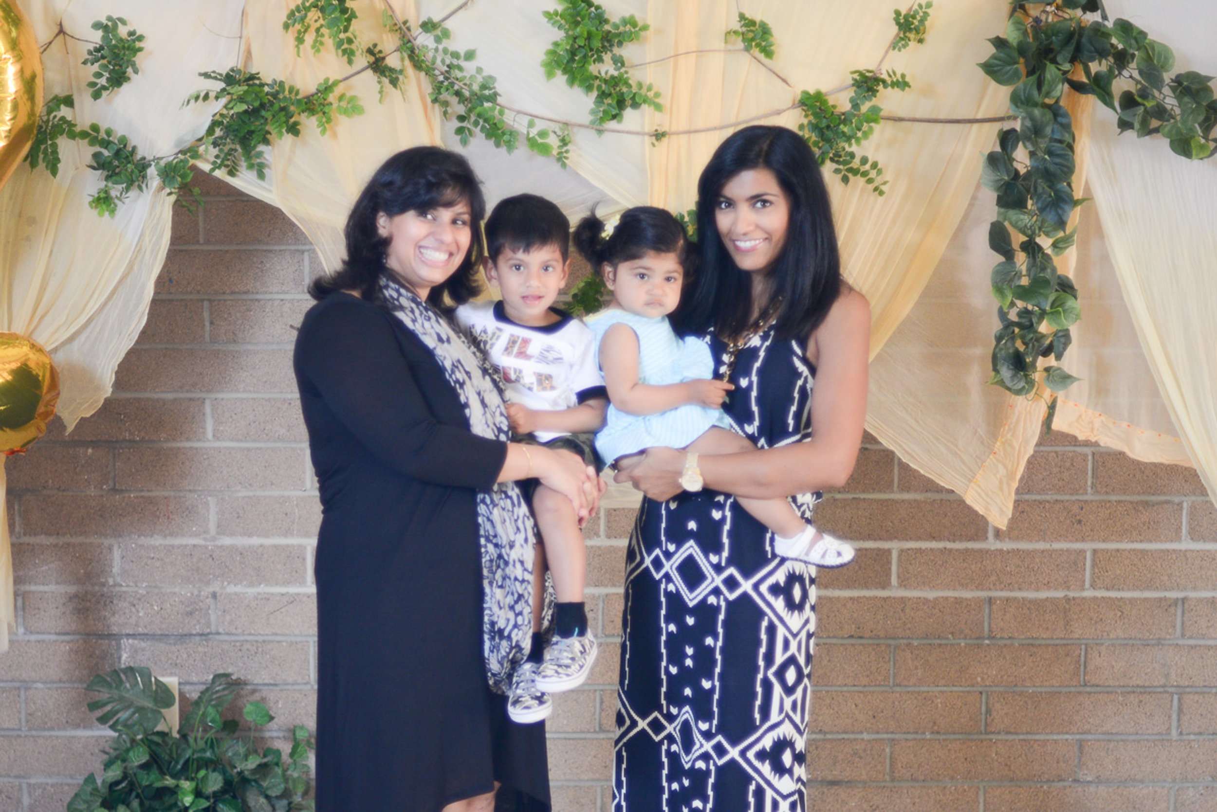 Event photography. Two women holding young children, standing in front of a decorated backdrop with greenery and ivory fabric drapes, against a brick wall.