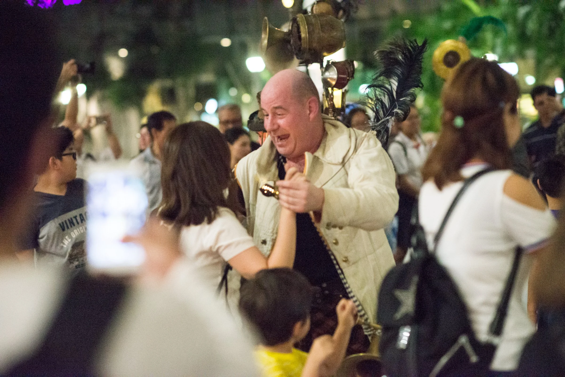 A bald man wearing a white jacket is smiling and holding hands with a young girl in a white shirt, surrounded by a crowd in an outdoor setting at night.
