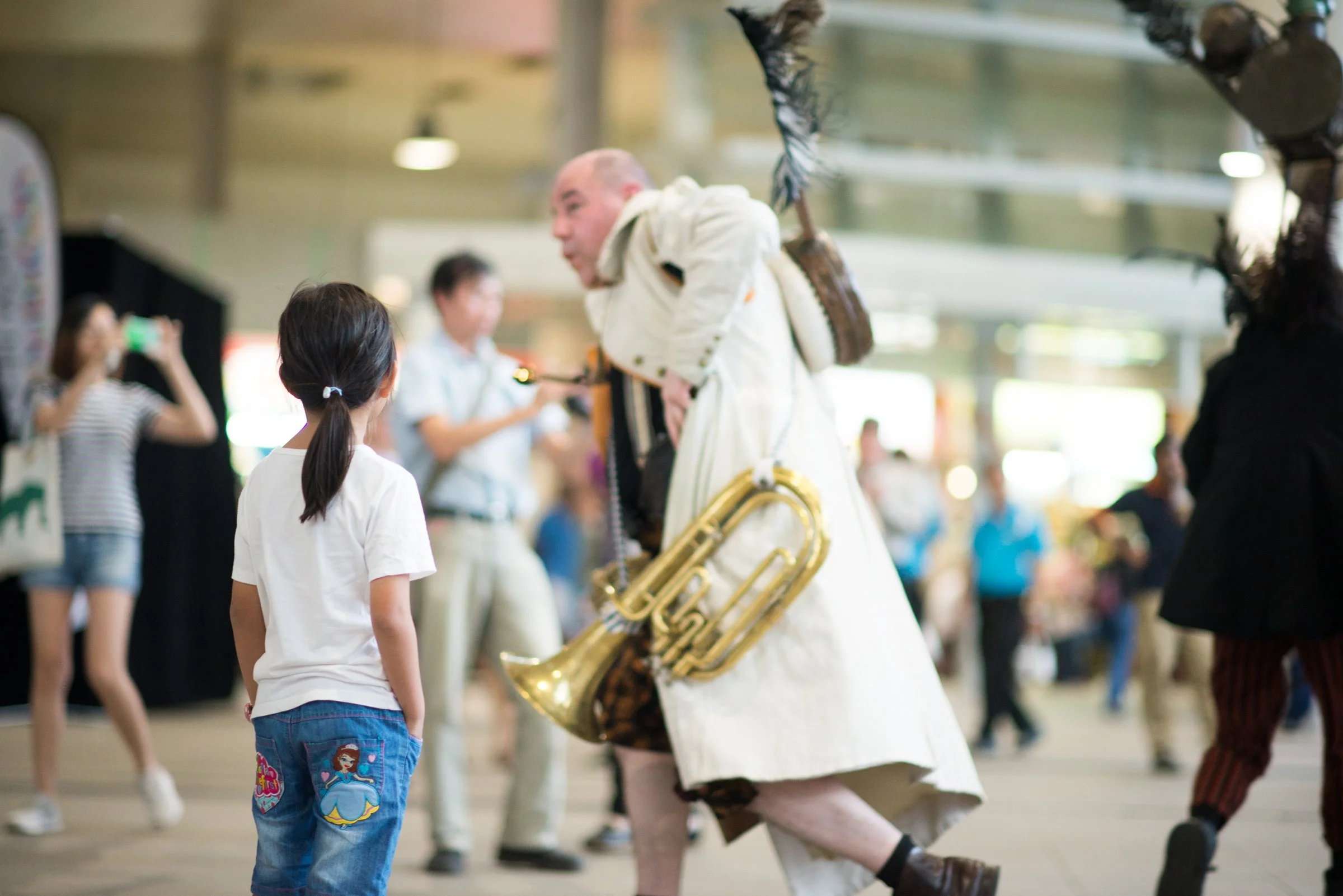 A street performer dressed in a white coat and hat, carrying a guitar and a brass instrument, interacts with a young girl at an indoor event.