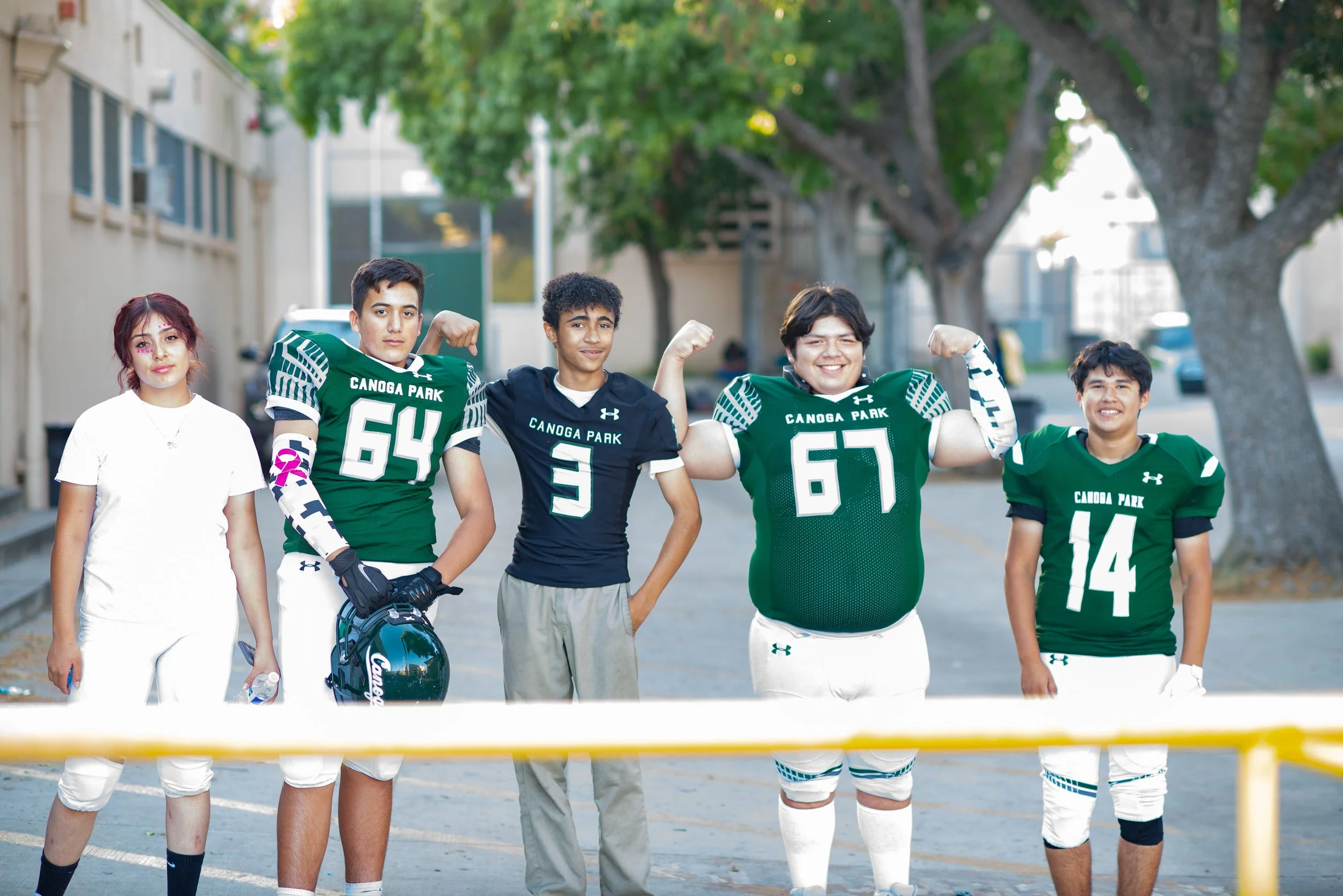 School photography. Group of five young people standing outdoors, with two wearing green football jerseys labeled Canoga Park, in a casual pose, and smiling, in front of trees and a building.