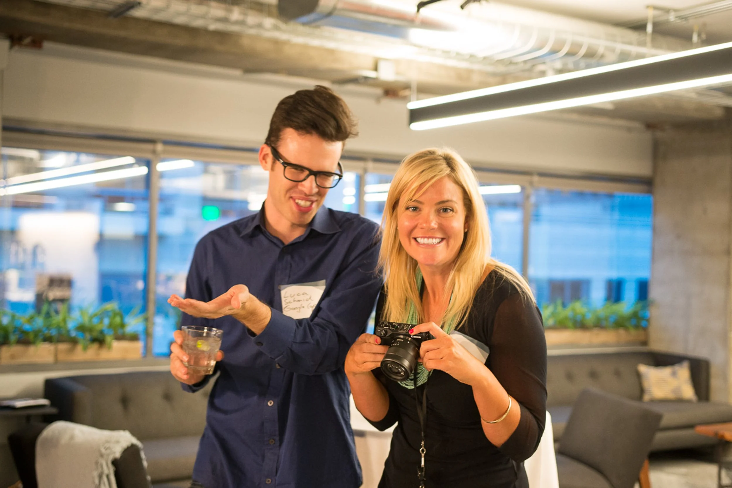 A young man and a woman smiling indoors, with the woman holding a camera and the man holding a glass of drink.
