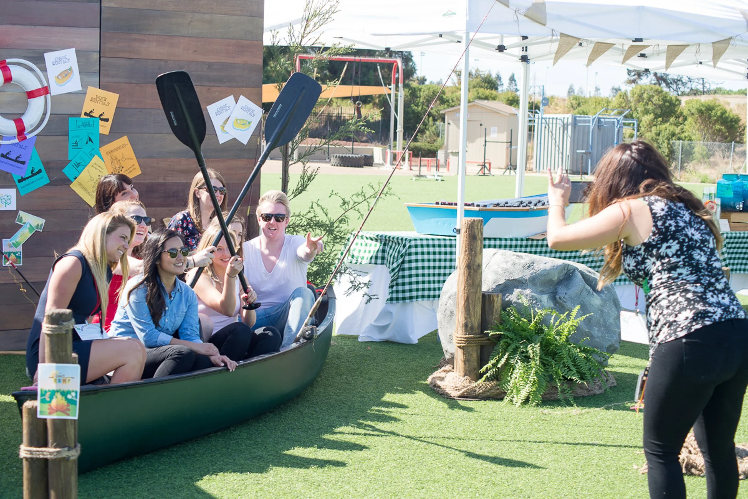 A group of women sitting inside a canoe, enjoying a boat-themed photo op, while a woman in front appears to be taking a picture of them, outdoors on a bright, sunny day at a festival or event with decorations and a tent in the background.