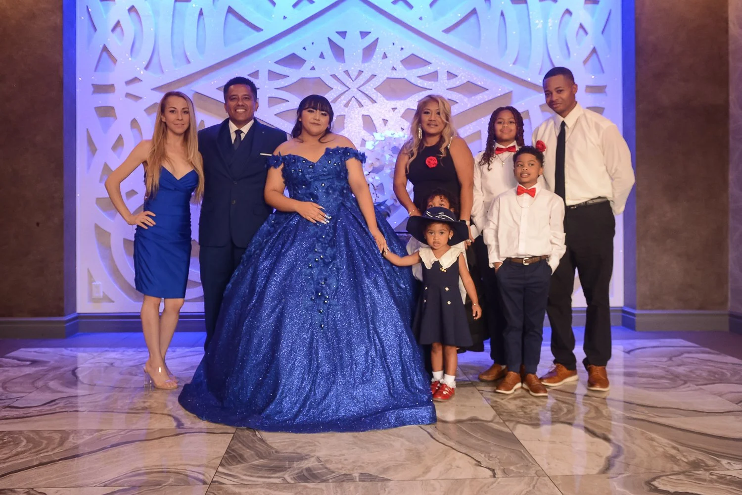 Event photography. Quinceañera. Family group photo with seven members dressed in formal attire, standing in front of a decorative wall with blue lighting, including women in dresses and children in shirts and dresses.