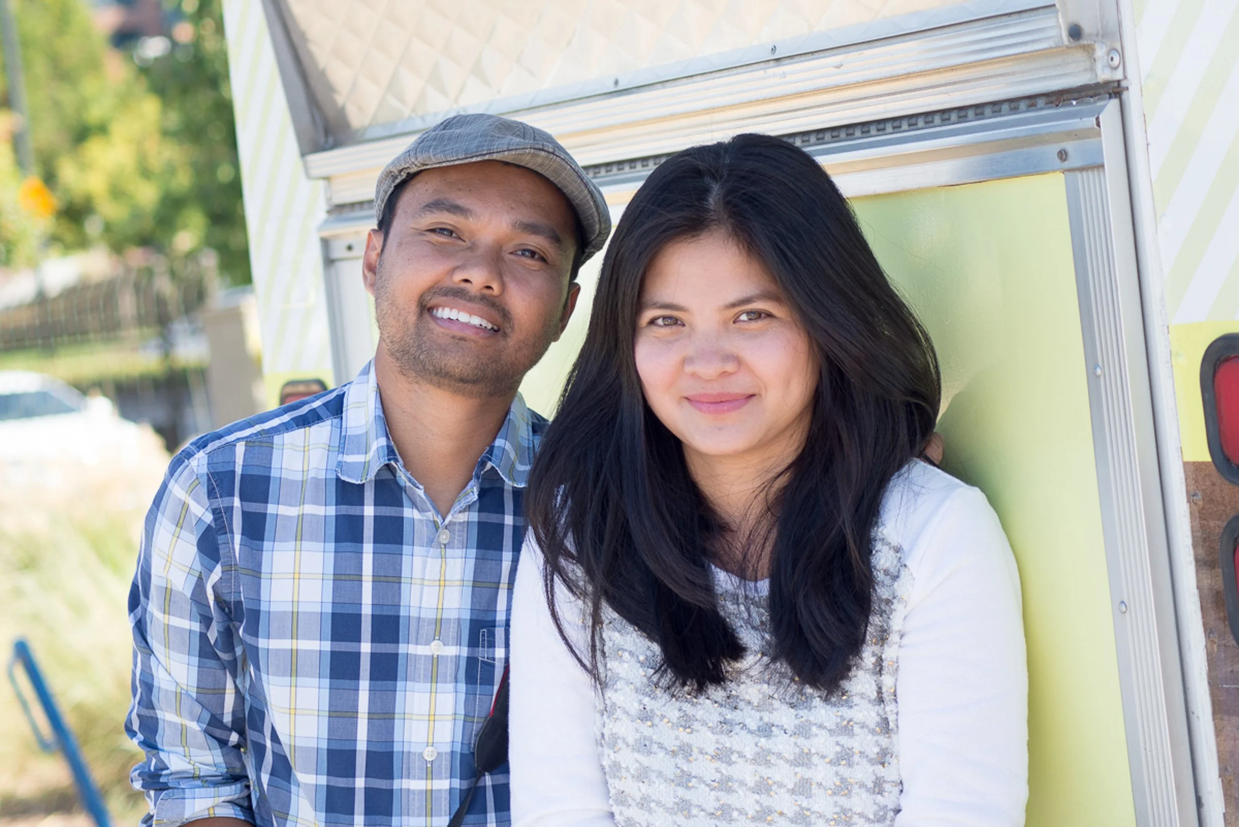 A smiling man and woman standing outdoors next to a food truck.